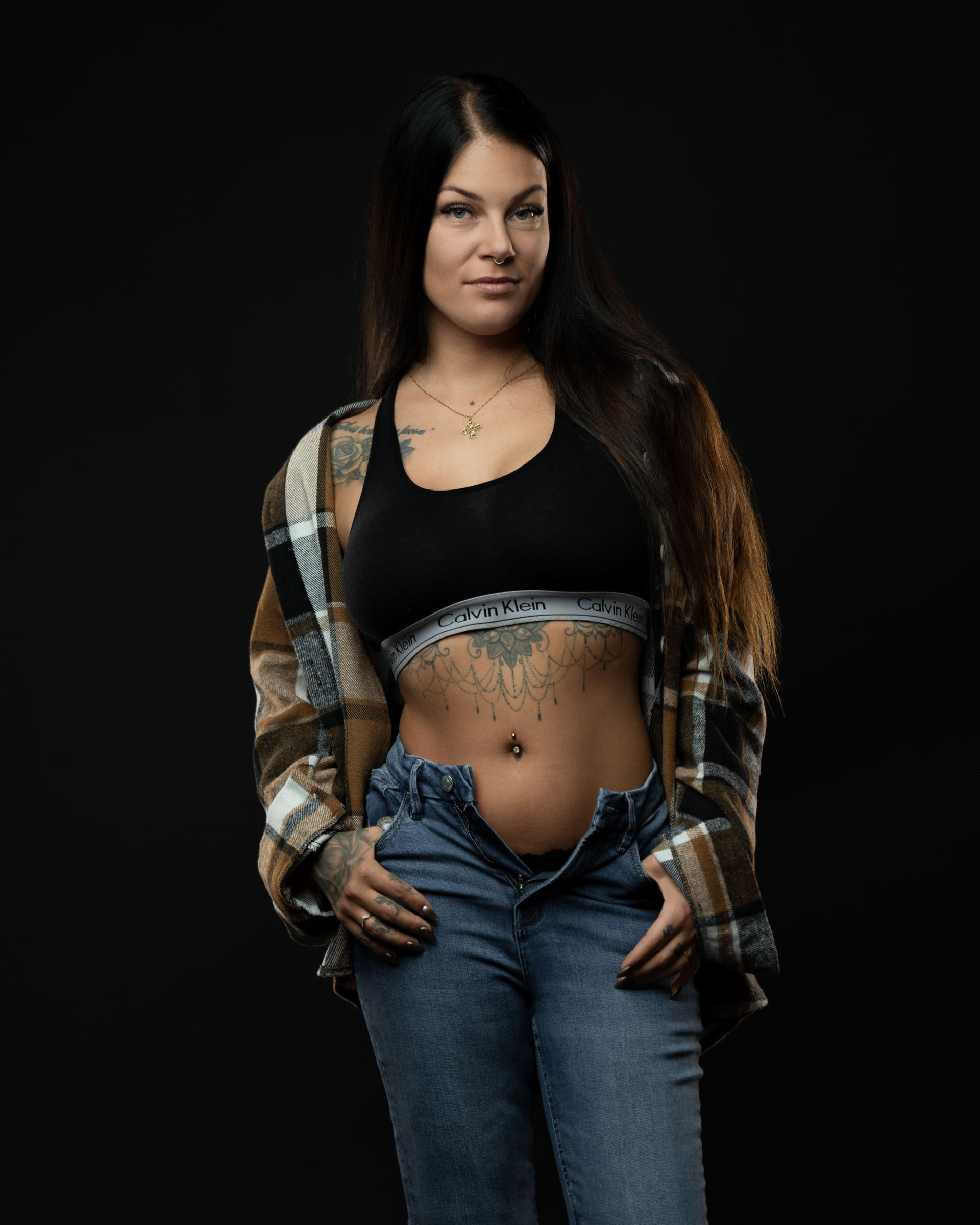 Studio portrait of Amanda wearing a black sports bra, open jeans and a plaid shirt, posing confidently against a dark background