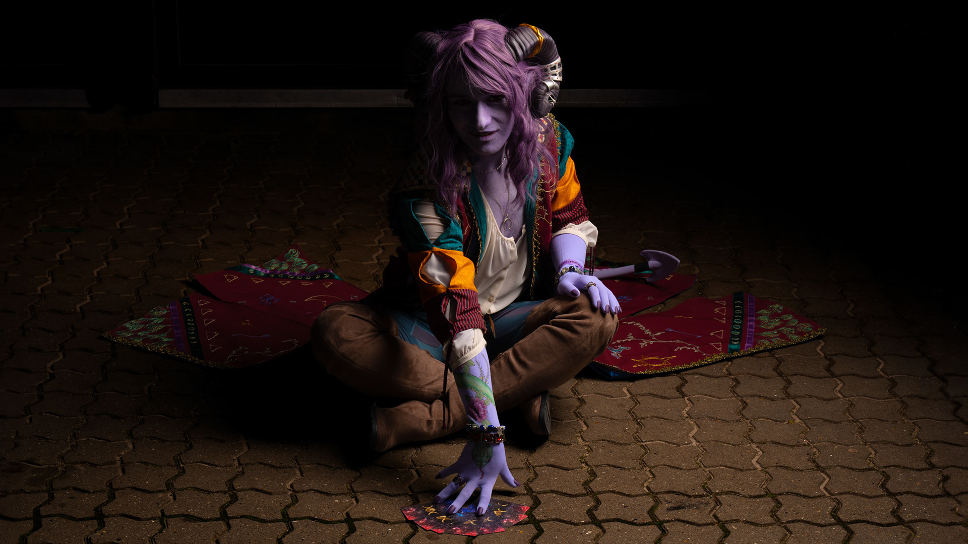Jester Lavorre cosplay kneeling on the ground, reaching toward a spread of tarot cards, purple skin and embroidered coat visible in the lighting.