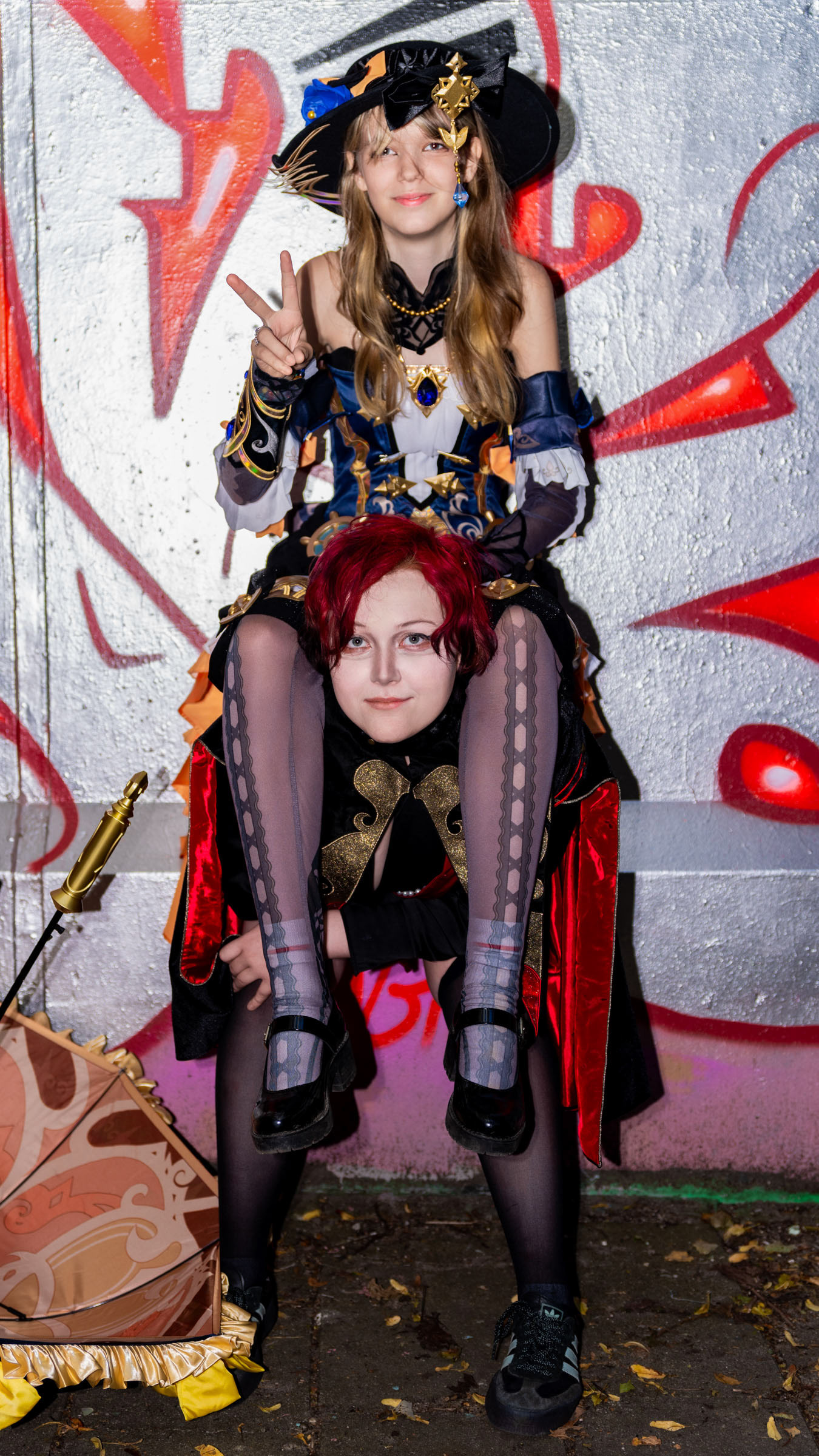 Cosplayer ruxqym sitting on the shoulders of cosplayer zen.c0z while making a peace sign, both in detailed costumes and posing in front of a bright graffiti-covered wall.