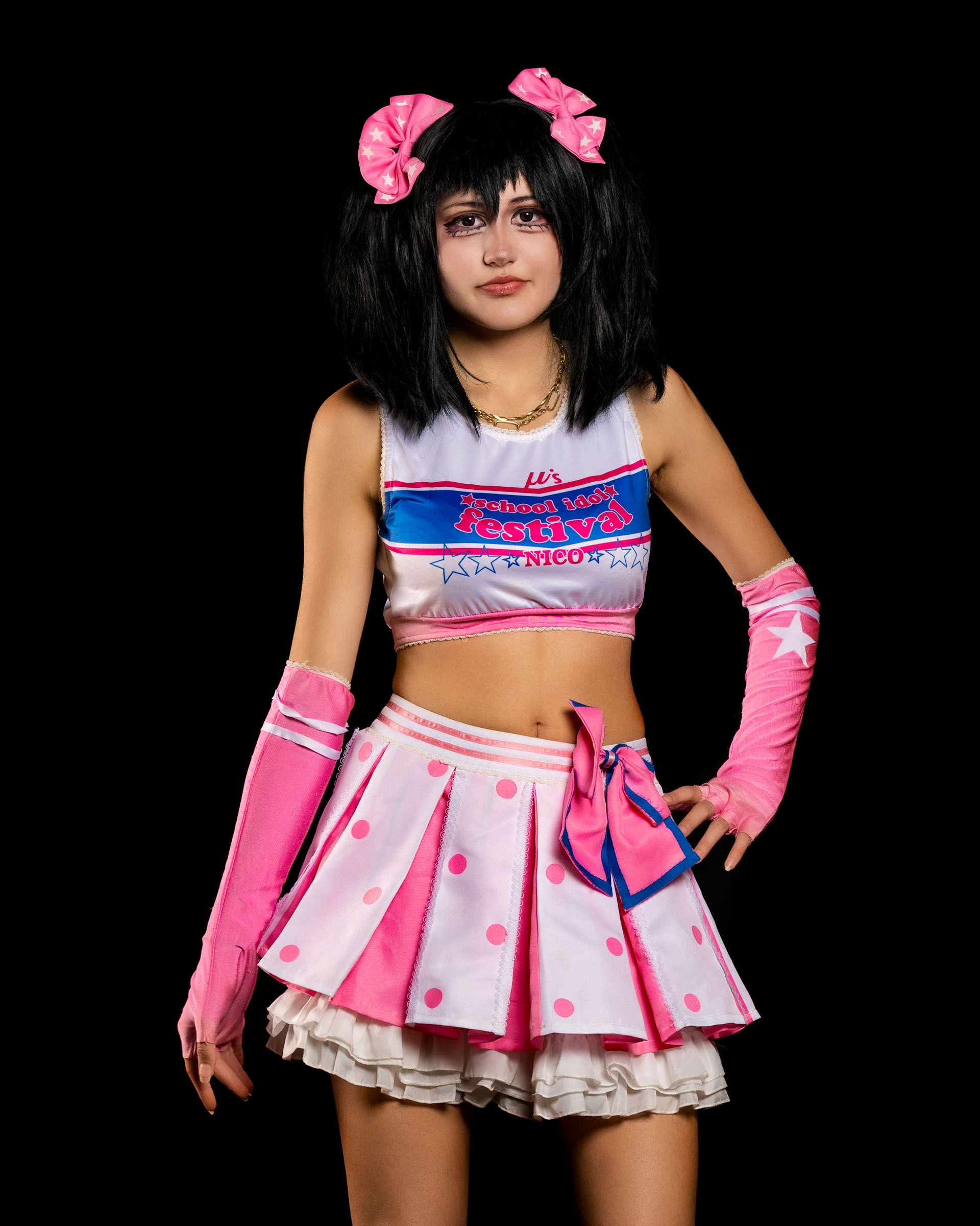 Full-body pose of may_c0z dressed as Nico Yazawa, standing confidently in the pink idol uniform against a black background.