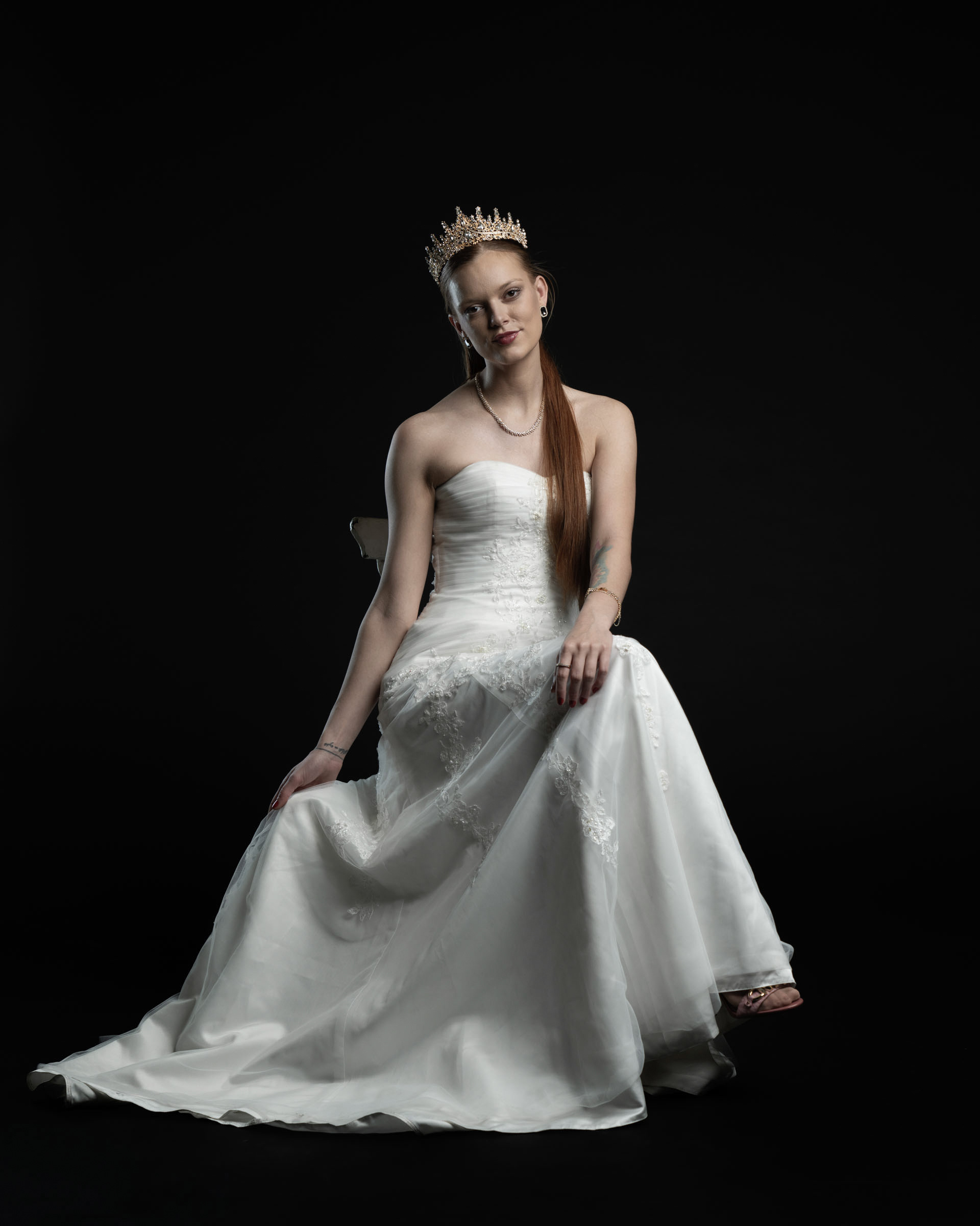 Model sitting in a white dress with crown, hands resting on her lap, posing calmly.
