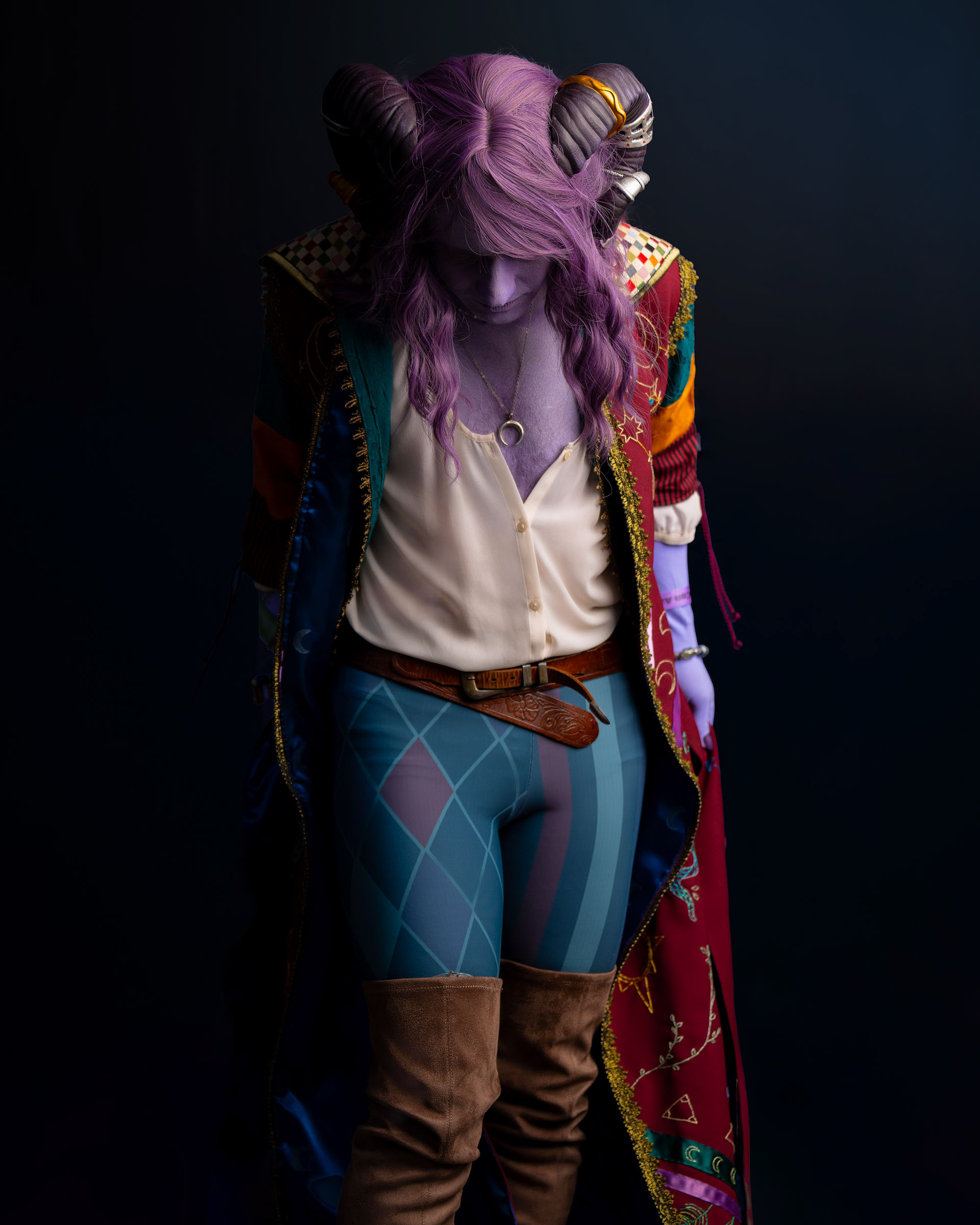 Cosplayer dressed as Jester Lavorre looking down, purple skin, curled horns and detailed embroidered coat, lit dramatically against a dark teal background.