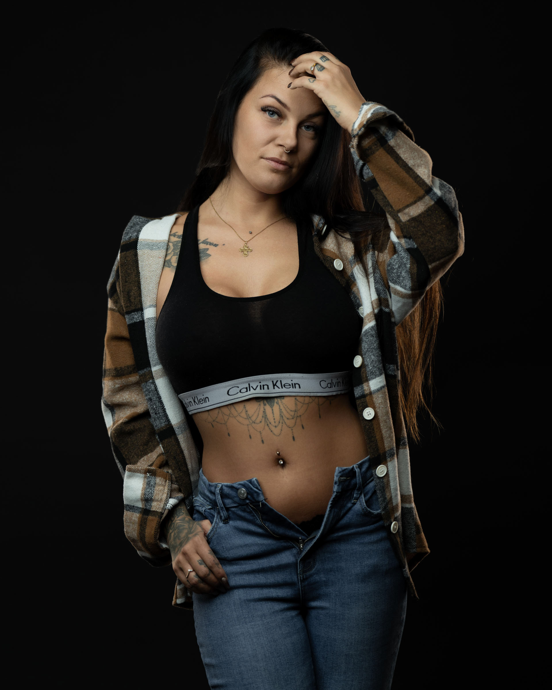 Amanda touching her hair while wearing a plaid shirt, black sports bra and jeans, photographed in a minimal dark studio.