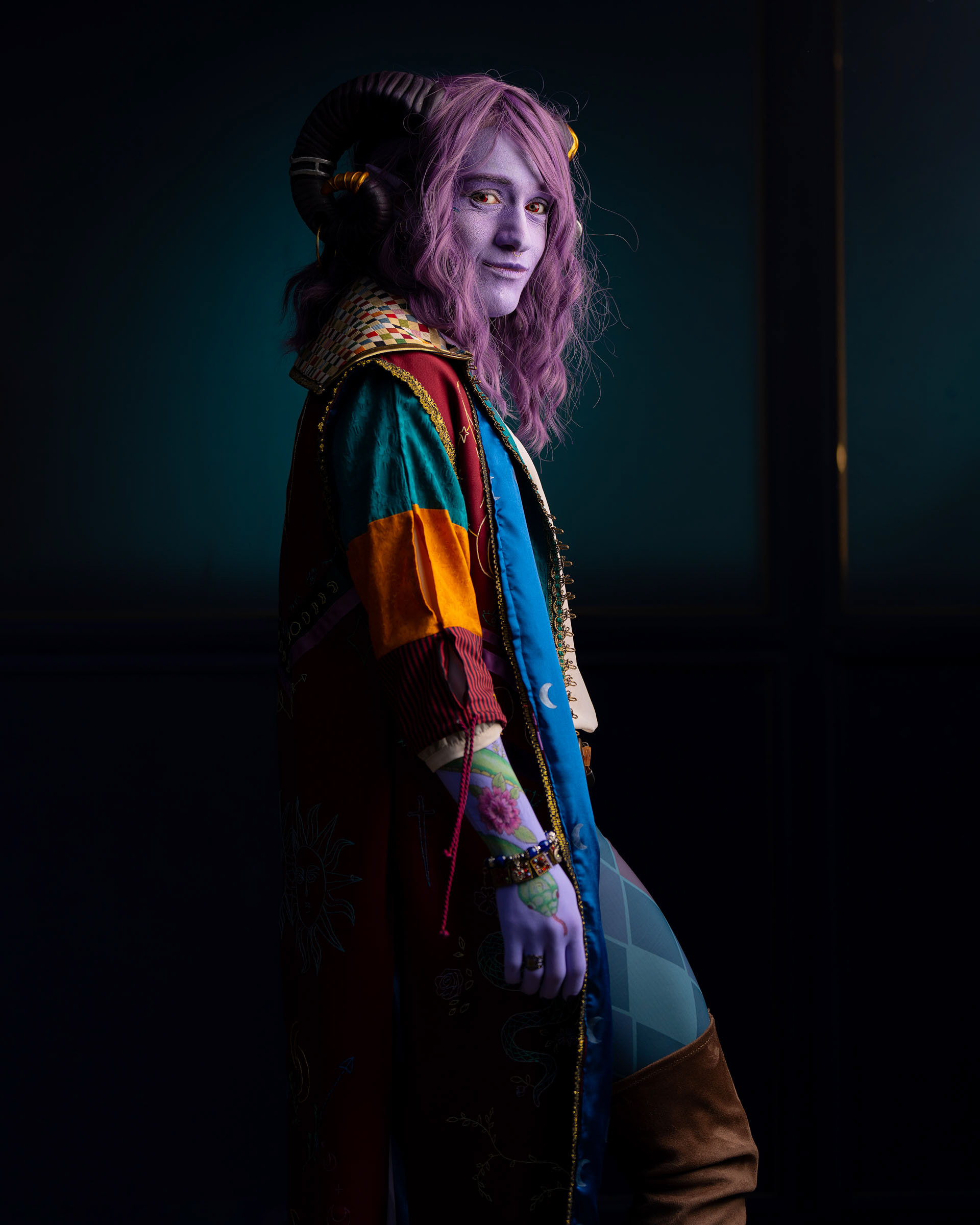 Side portrait of Jester Lavorre cosplay with purple skin, violet hair, curled horns and a vibrant multicolored coat, turning toward the camera.