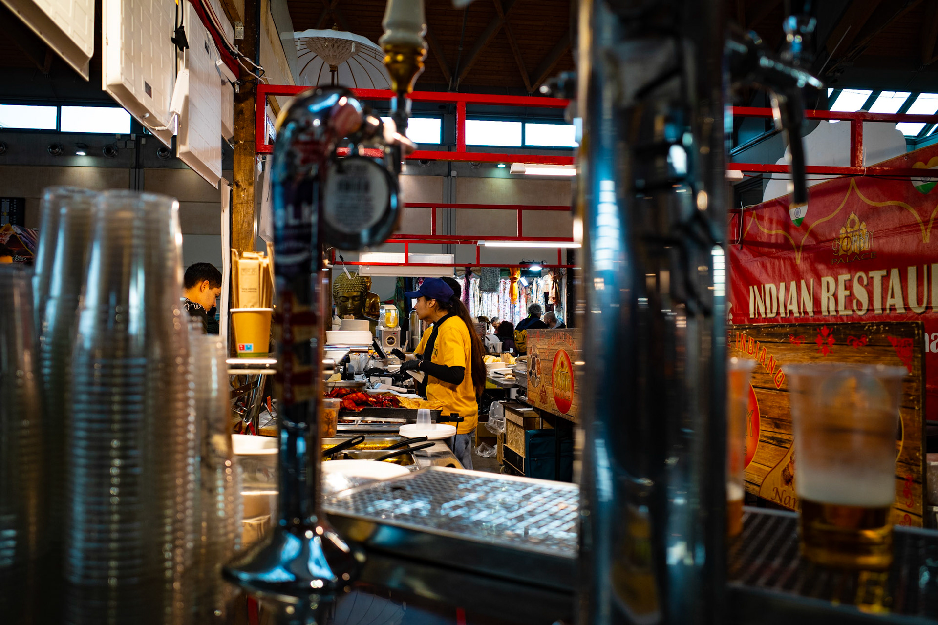 Between stacked glasses, metal taps and reflective surfaces, the gaze travels through a narrow improvised passage leading to a kitchen alive with movement. Quick hands prepare dishes while the murmur of the market fills the air. Warm colors, red signs and invisible aromas evoke distant journeys and traditions carried into the city. In this confined space, among tools and ingredients, the act of cooking becomes an everyday gesture connecting different cultures within the same urban rhythm.