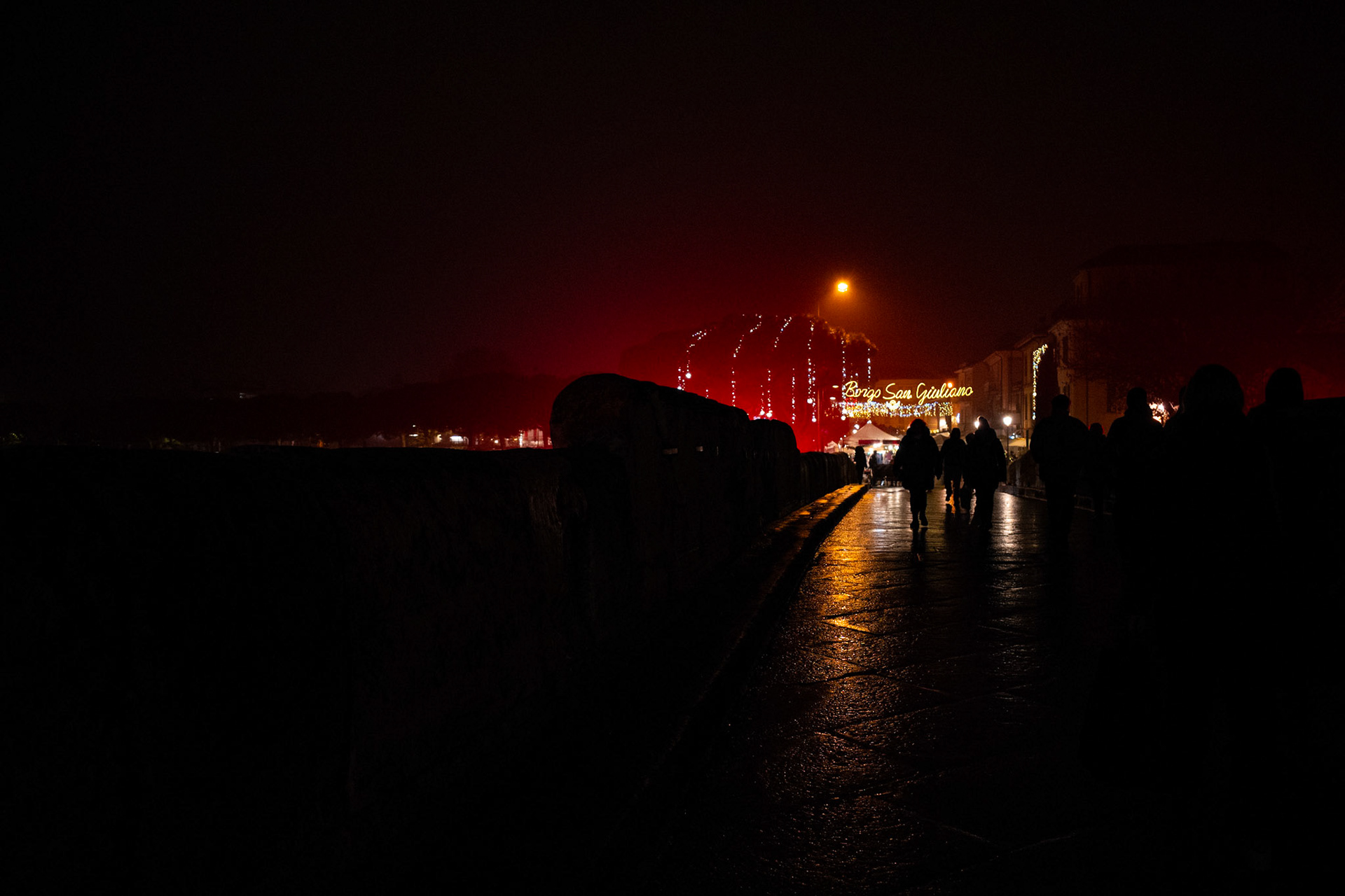 The wet road reflects an invitation that asks for no answer. Figures move like evening thoughts, suspended between the desire to arrive and the urge to remain at the edge. The celebration exists, but it does not claim the center: it is a distant call, while the night protects those who choose to stay in the dark.
