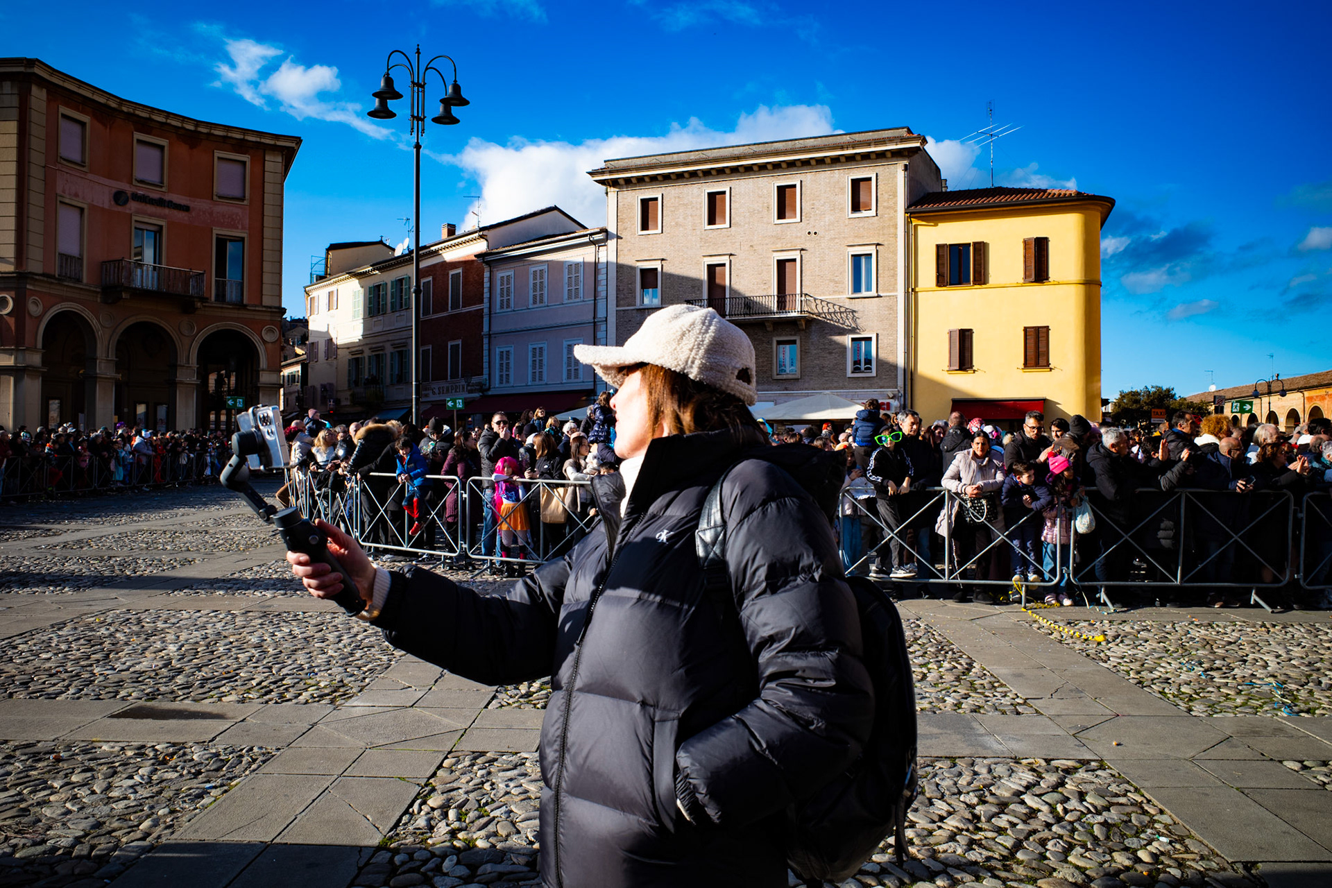 The square is filled with voices and anticipation, yet in front of the crowd one figure pauses and raises the phone like a small window into the present. The gesture is simple and ordinary, yet it turns the event into immediate memory. Behind the barriers the community watches the spectacle, while the screen gathers a personal version of the same moment. In that instant reality splits between experience and recording.