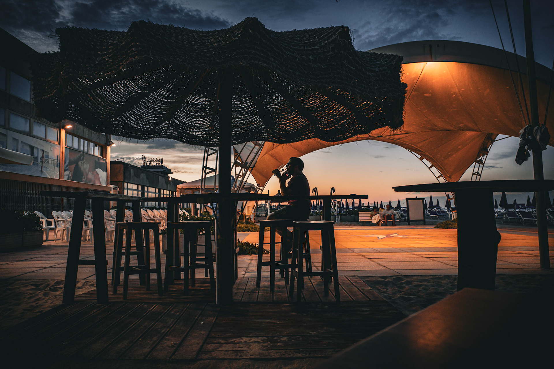 Between the distant sound of waves and the faint clink of glass, there is a silence that is not absence but suspension. A solitary figure remains still beneath woven shadows as the sky slowly fades beyond the horizon. Warm light rests on surfaces like a held breath, turning wood and air into something almost tangible. Empty chairs hold unspoken words. In this fragile space between sea and transparency, time dissolves, leaving only waiting behind.