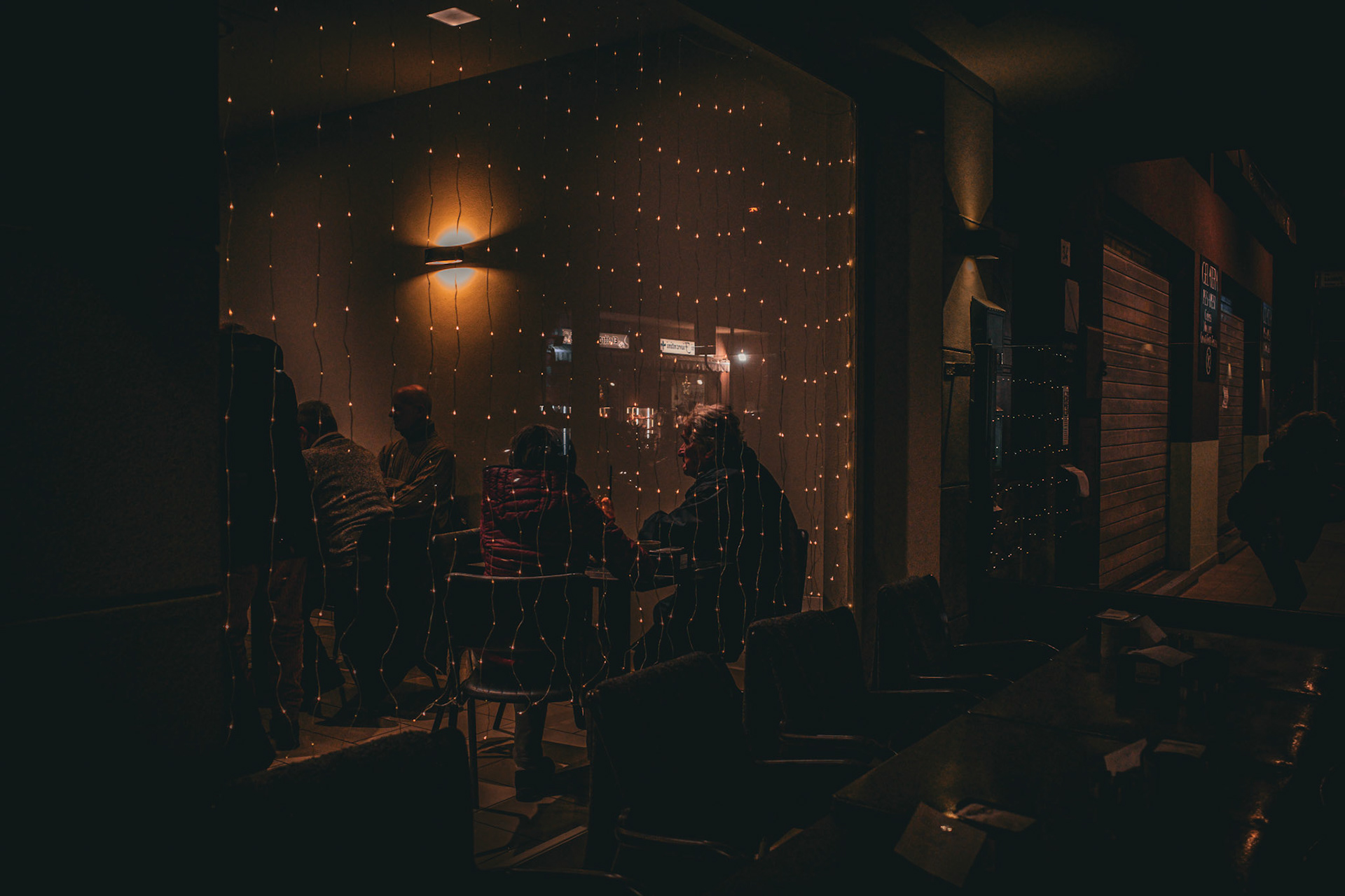 Behind the curtain of lights, the night protects slow gestures and voices that never reach the ears, only the gaze. The seated figures turn into gentle silhouettes, gathered around a warmth that cannot be seen, only felt. It is a suspended fragment, a place where quiet becomes companionship.