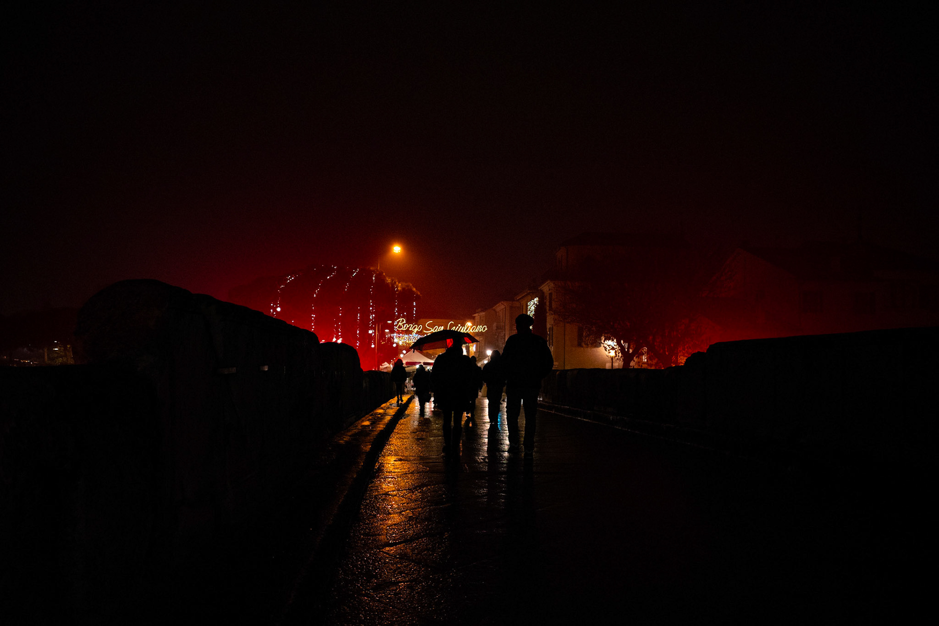 Dark figures move forward across a rain-polished bridge, while red light glows over what remains distant. Night wraps everything, turning the walk into a collective and silent gesture. There is no urgency, only the slow acceptance of a direction quietly shared.