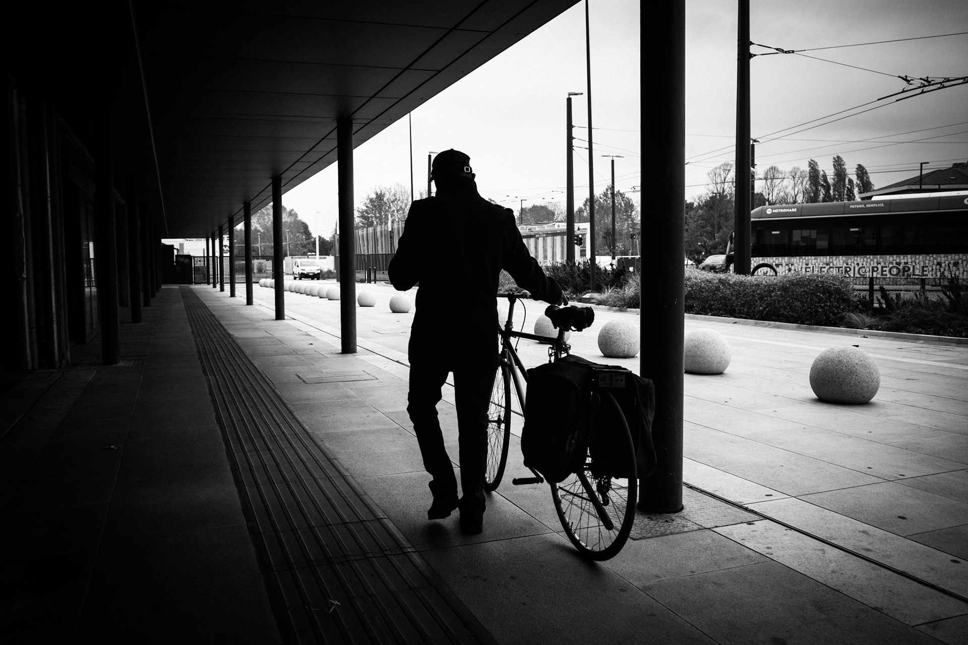 A shadow moves beneath the walkway, carrying the unhurried rhythm of a departure with no urgency. The world around dissolves into lines, pillars and wind, while the bicycle stands as a silent companion. In this frame of greys and distance, every step becomes a thought taking shape.