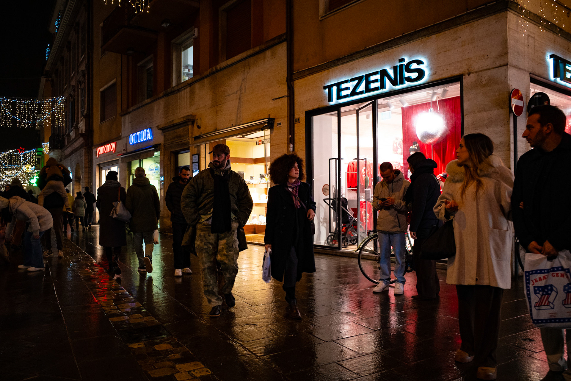 The glossy street reflects lit shop windows and moving faces, while the evening wraps every gesture in apparent calm. People flow like distracted thoughts, suspended between purchases, waiting, and brief encounters. In this urban theatre, light does more than illuminate: it reveals fragments of humanity in transit.