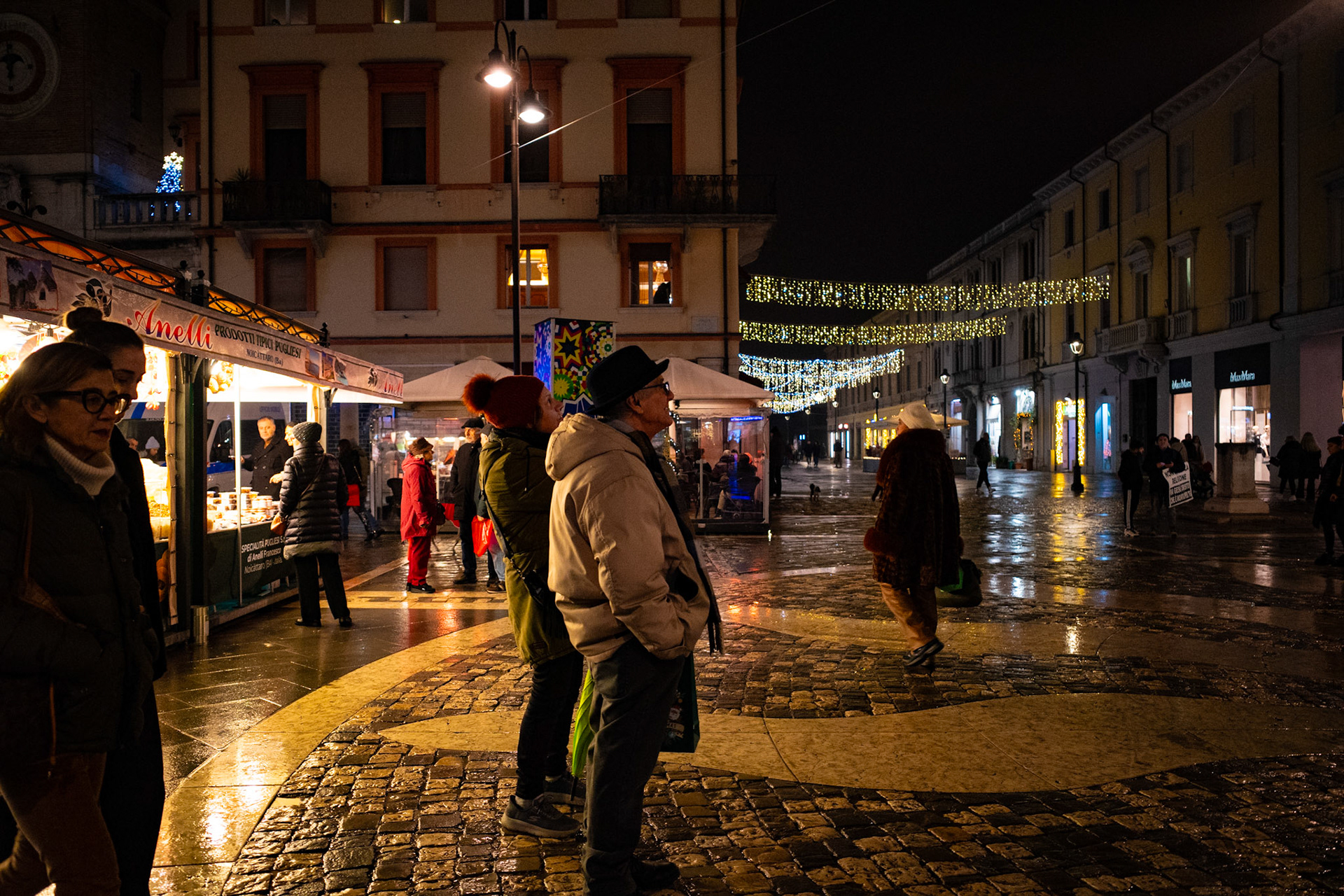Rain turns stone into a mirror and the city into a silent stage. Lights trace fleeting paths above bowed heads and listening gazes. Every figure is waiting for something, even just a moment, while the night moves slowly and carries the weight of footsteps.