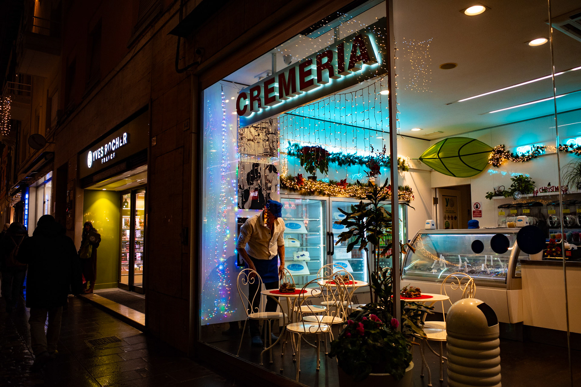 Behind the glass, light breathes like a warm heart, while outside the street flows cold and unaware. A simple gesture arranges chairs and silences, turning an urban corner into a temporary shelter. There, the night slows down, watching humanity reflect between artificial lights and everyday desires.