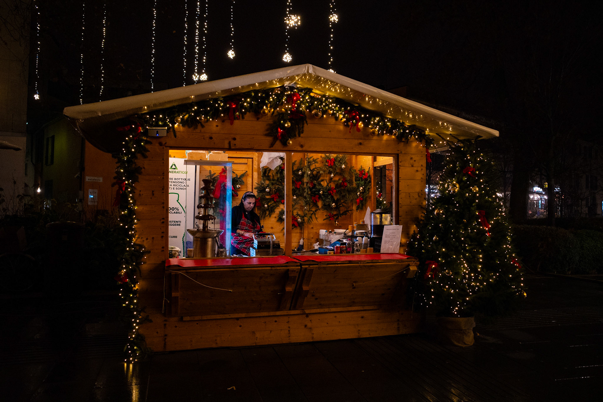 In the heart of the urban night, a lit wooden cabin becomes shelter and boundary. Lights trace a warm perimeter, wood speaks of patient waiting, while an everyday gesture unfolds calmly. It is a temporary place, yet essential, where the city slows down and grants itself a quiet truce.