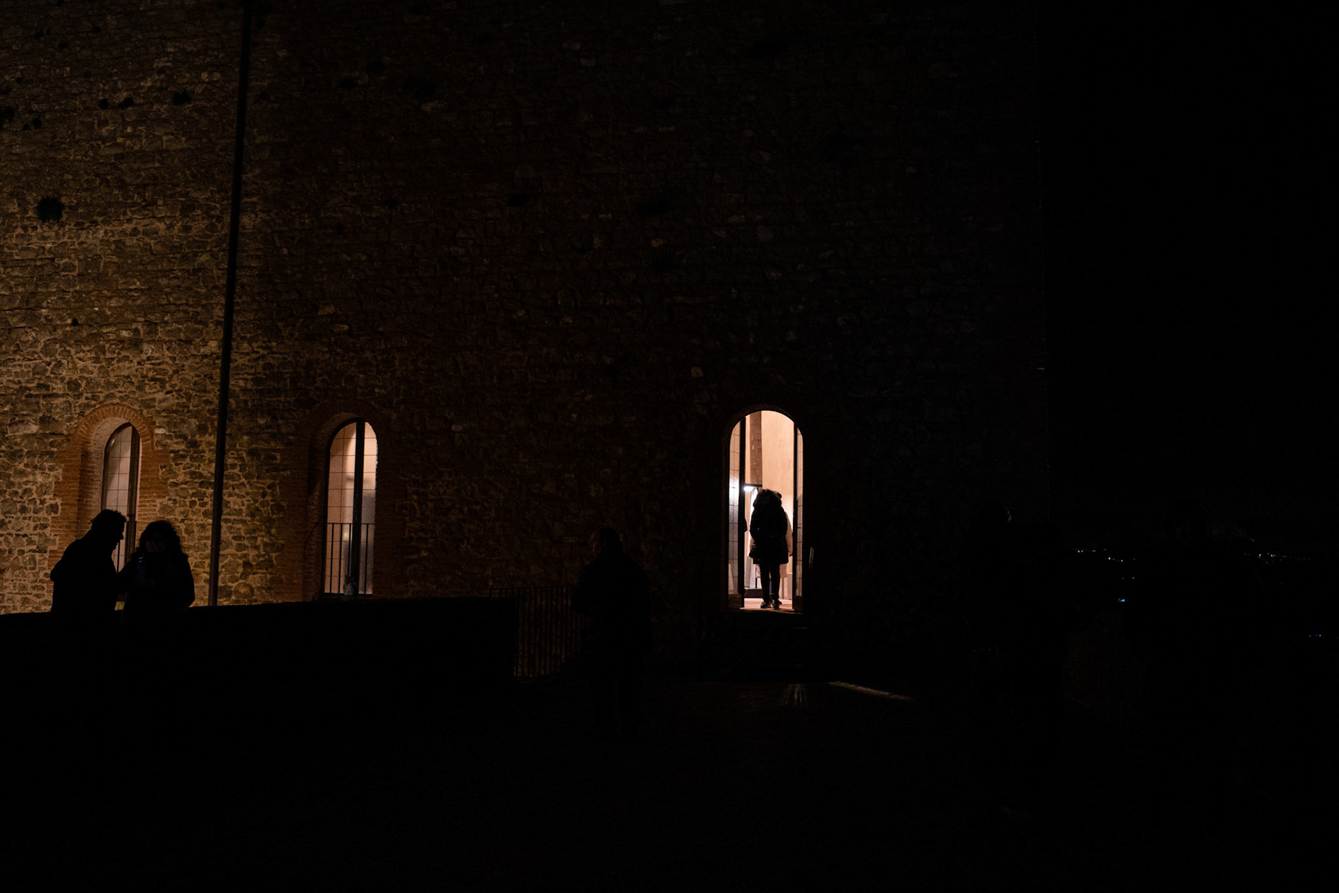 Stone absorbs the silence and releases it as slow-moving shadows. A warm light seeps through the openings like a fragile invitation, while figures hesitate between inside and outside. The night does not observe: it waits. And every crossing becomes an intimate act, almost sacred.