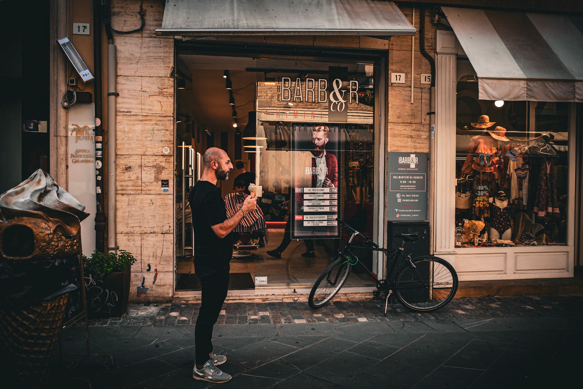 He stands just outside the doorway, holding something simple in his hand, while reflections multiply behind the glass. Inside, mirrors repeat gestures, faces, fragments of routine. Outside, the street continues its indifferent rhythm. The pause is brief but deliberate, a thin line between movement and stillness. In front of mirrors that reflect more than appearances, the moment becomes suspended, as if even the city needed a breath before continuing.