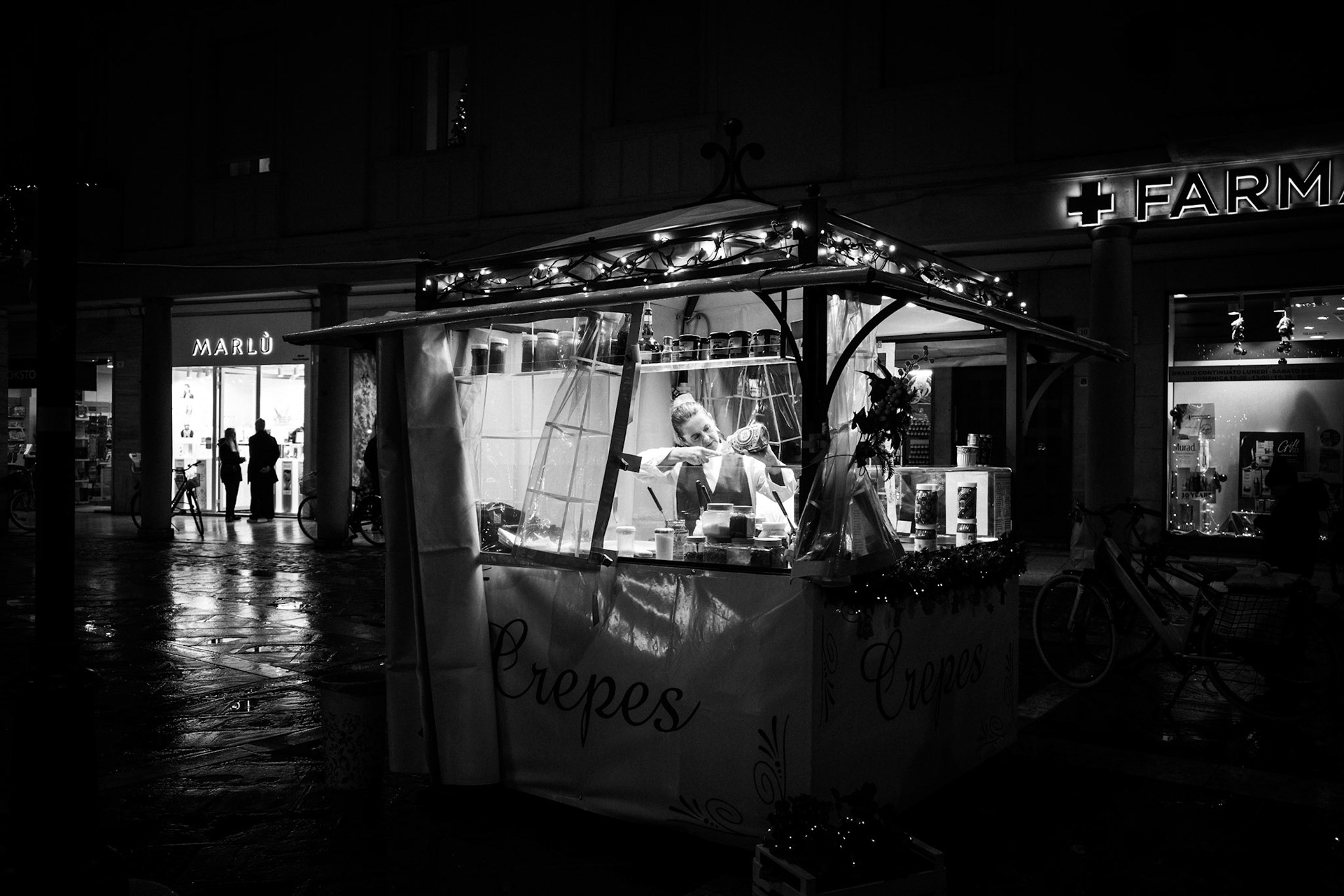 Rain polishes the city like a memory just born. A small refuge of light floats in the darkness, while patient hands repeat ancient, necessary gestures. Outside, everything flows on footsteps, shadows, indifferent shop windows. Inside, time slows down and becomes human. It is in these fragile spaces that the night stops being empty and learns how to listen.