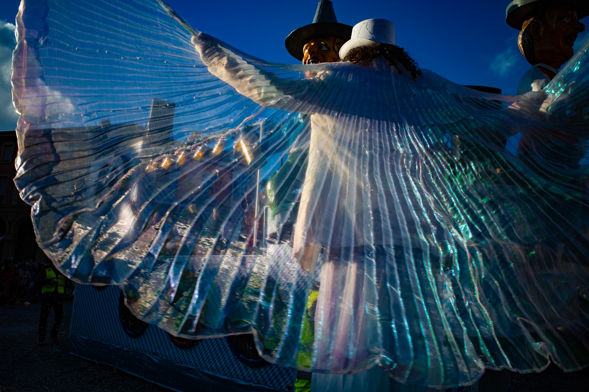 Facing away from the crowd, a figure spreads translucent wings like crystalline membranes pierced by sunlight. The sky becomes a dramatic abyss of blue, while playful shapes and bold colors erupt around the scene. It is not an escape, but a quiet act of presence. The body turns into a reflective surface, absorbing and returning light. Among masks, floats and curious eyes, a collective ritual unfolds where childhood and dream walk beside matter.