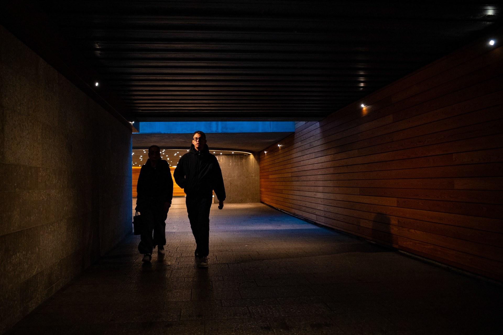 Two figures emerge from the depth of a narrow passage, their steps measured and their silhouettes carved by contrast. Darkness presses in from the walls, while a thin blade of cold light ahead suggests an exit, or perhaps only another threshold. The silence feels dense, almost tangible, as if the corridor itself were holding its breath. Between shadow and glow, the walk becomes more than movement: it turns into a quiet search for direction, for clarity, for that final fragment of light that still refuses to fade.