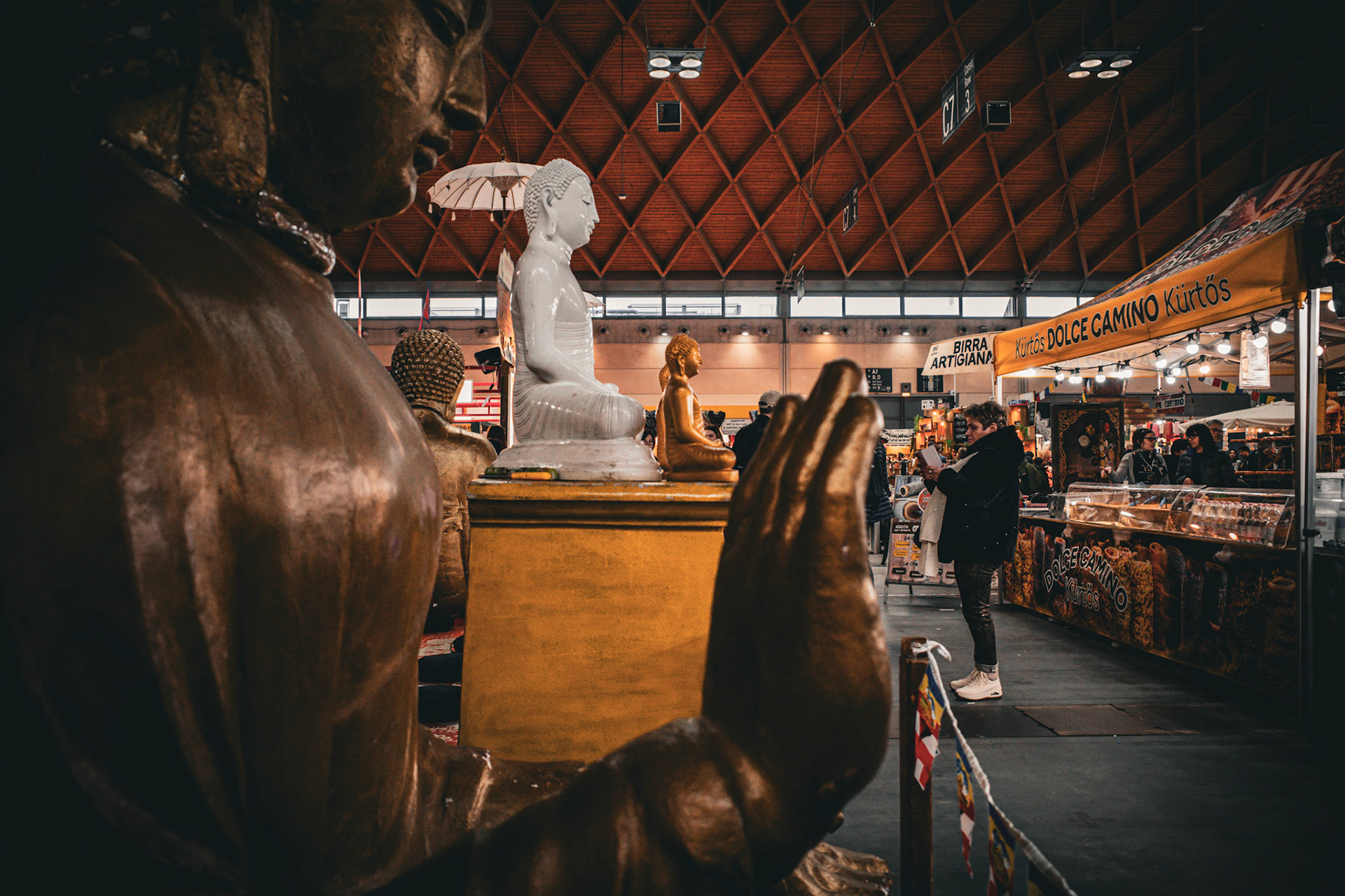 Between still statues and glowing stalls, an ancient gesture remains suspended. A raised hand watches the passing flow while trade keeps breathing. The sacred does not interrupt: it accompanies. In this shared space, silence finds room beside desire and makes it, for a moment, more human.