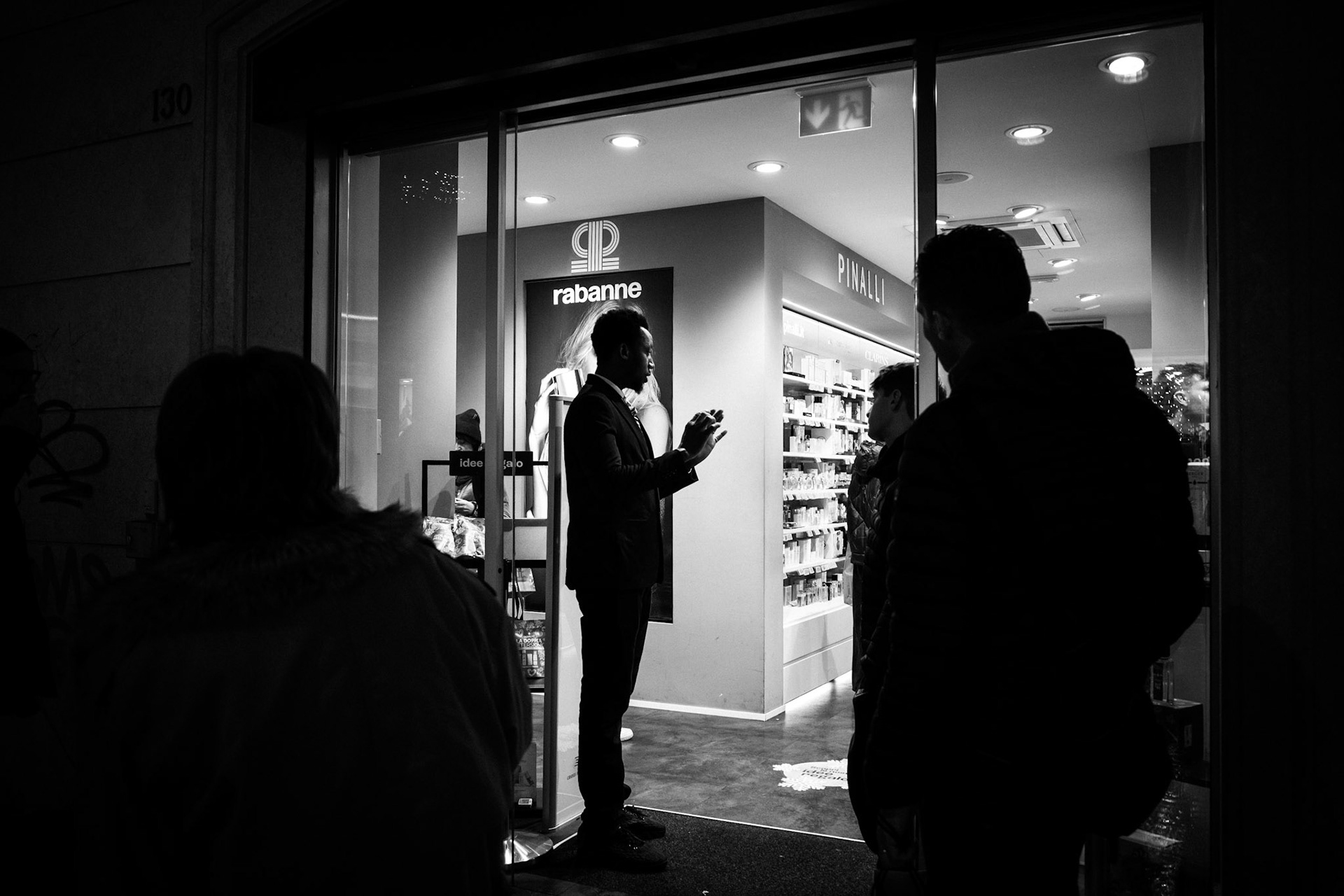 The interior light defines the gesture and separates it from the night. A man speaks with his hands, weighing words in the air, while surrounding figures listen without stepping forward. It is a suspended moment, where the threshold becomes a place of exchange and the crossing remains on hold.
