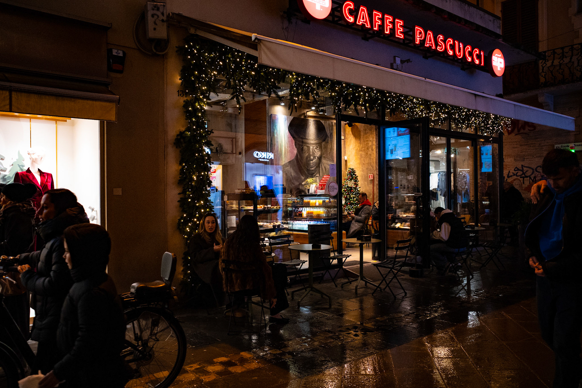Behind the glass, a painted face watches the flow of the street without taking part. Passersby move, speak, stop for coffee, while that gaze remains fixed and silent. It is a still presence within continuous motion, a witness that does not ask for attention but quietly holds it.
