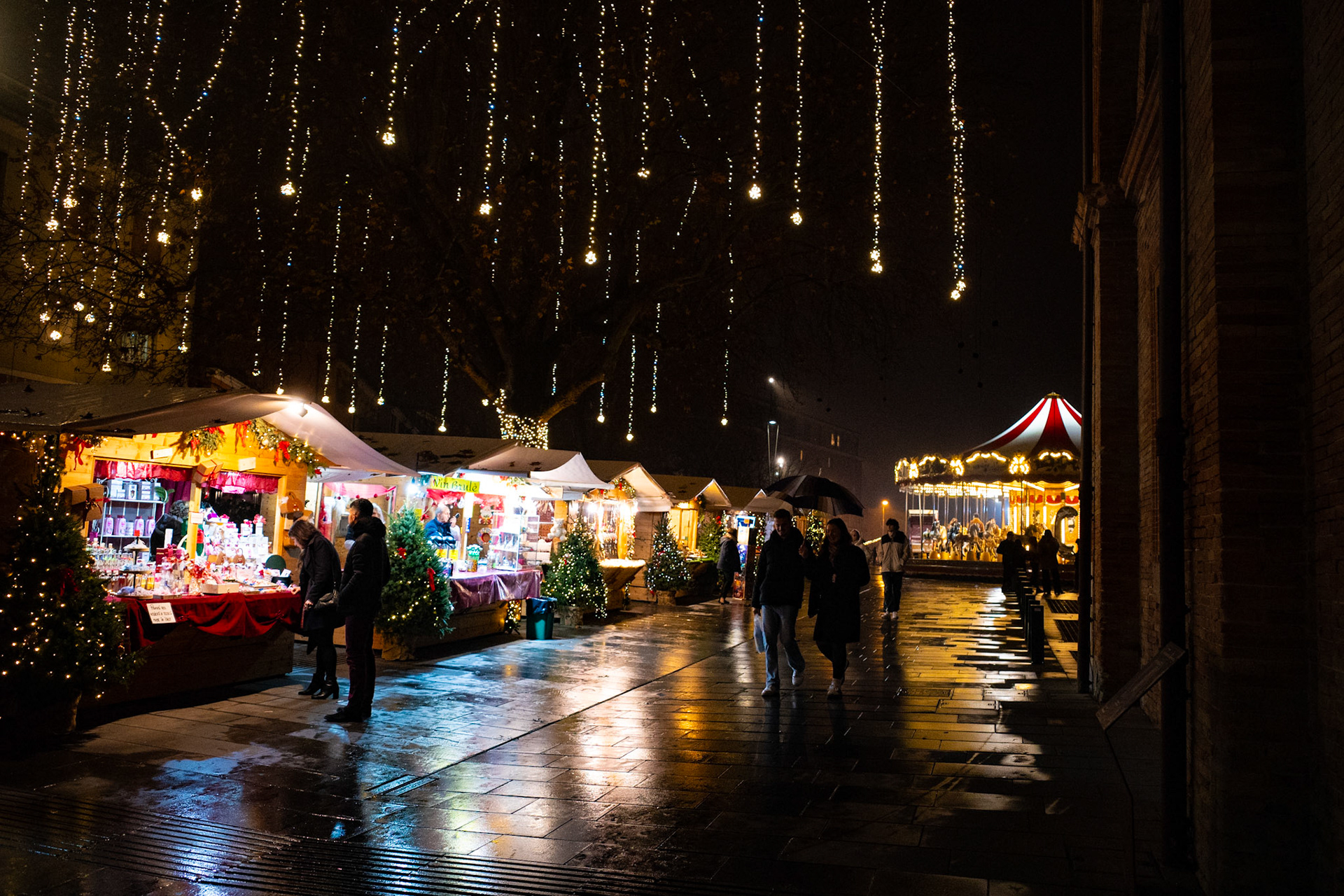 Lights fall from the trees like patient stars, reflecting on the wet pavement. The stalls glow, voices blend with the slow steps of passersby. It is a night that does not rush, inviting people to stay, where the city wears the fragile face of celebration.
