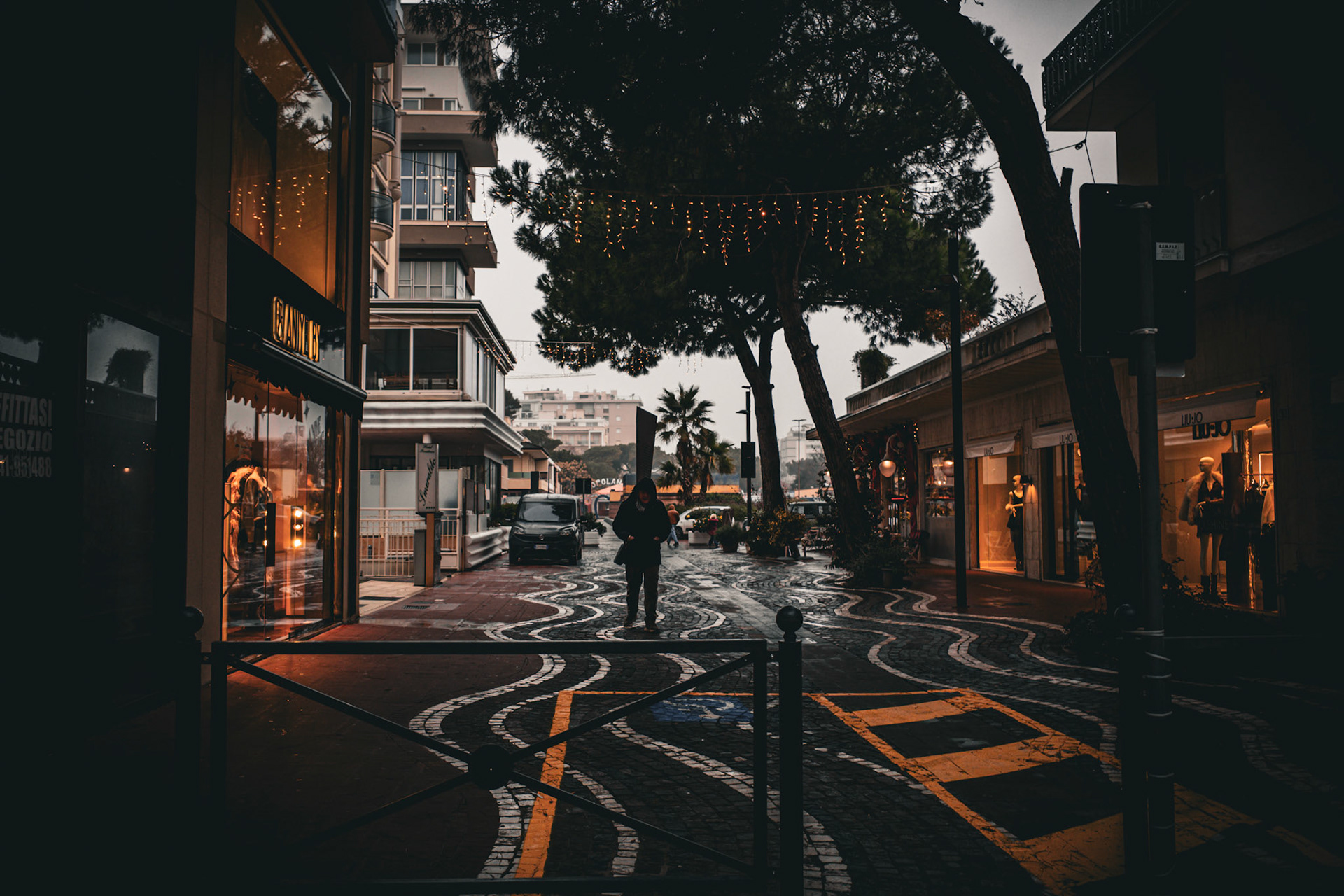White lines flow across the asphalt like marks left by an unseen hand. A figure moves forward at the center, suspended between lit windows and still trees. It is a quiet crossing, where the city guides footsteps and time waits.