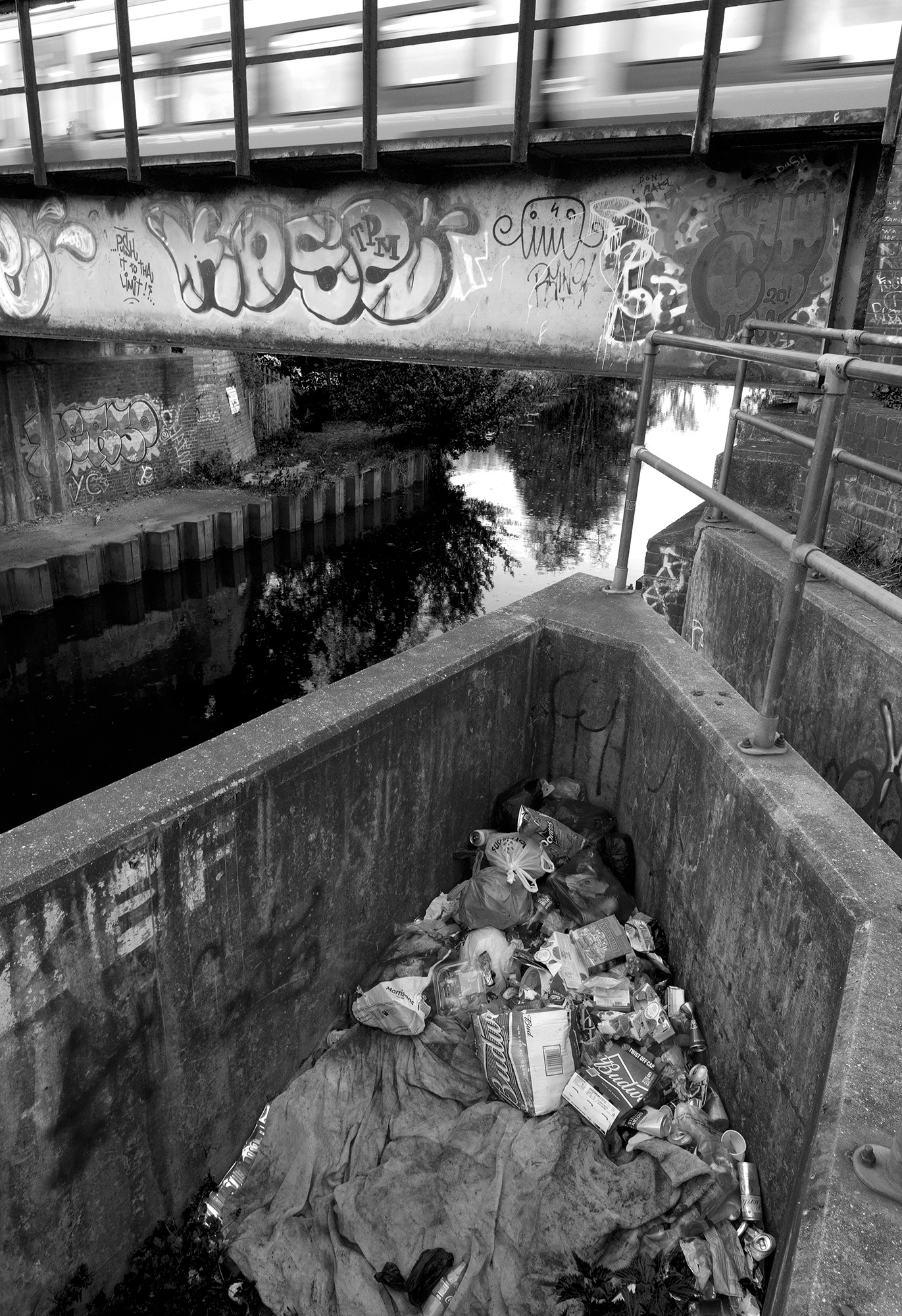 A black and white photograph of a river spanned by a bridge covered with graffiti with a large pile of rubbish in the foreground
