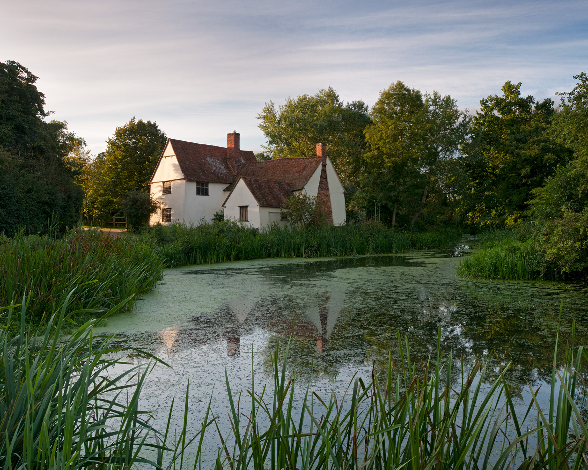 A colour photo of an old cottage reflected in a reed-fringed pool early on a summer morning