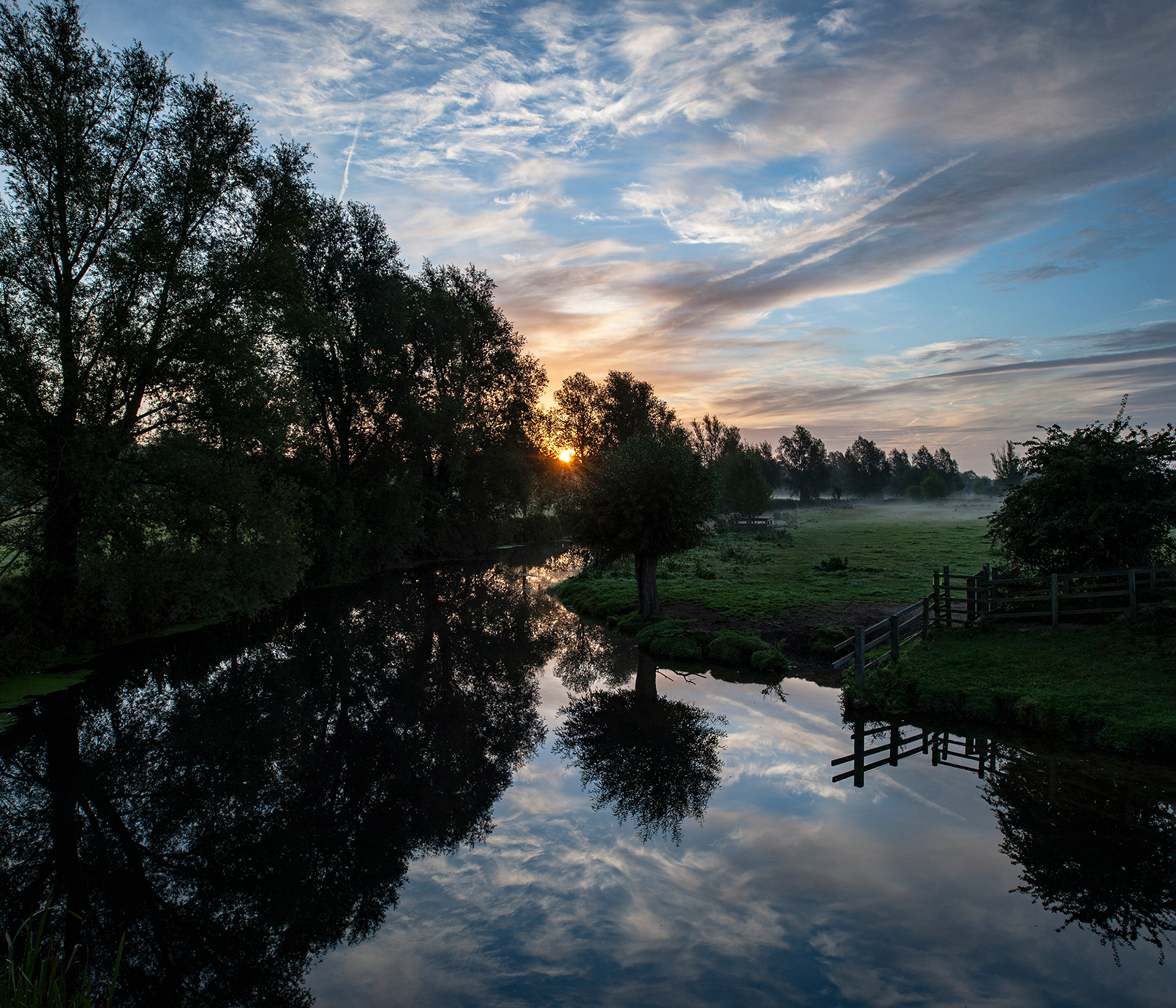 A colour photo of trees reflected in a tranquil river at sunrise