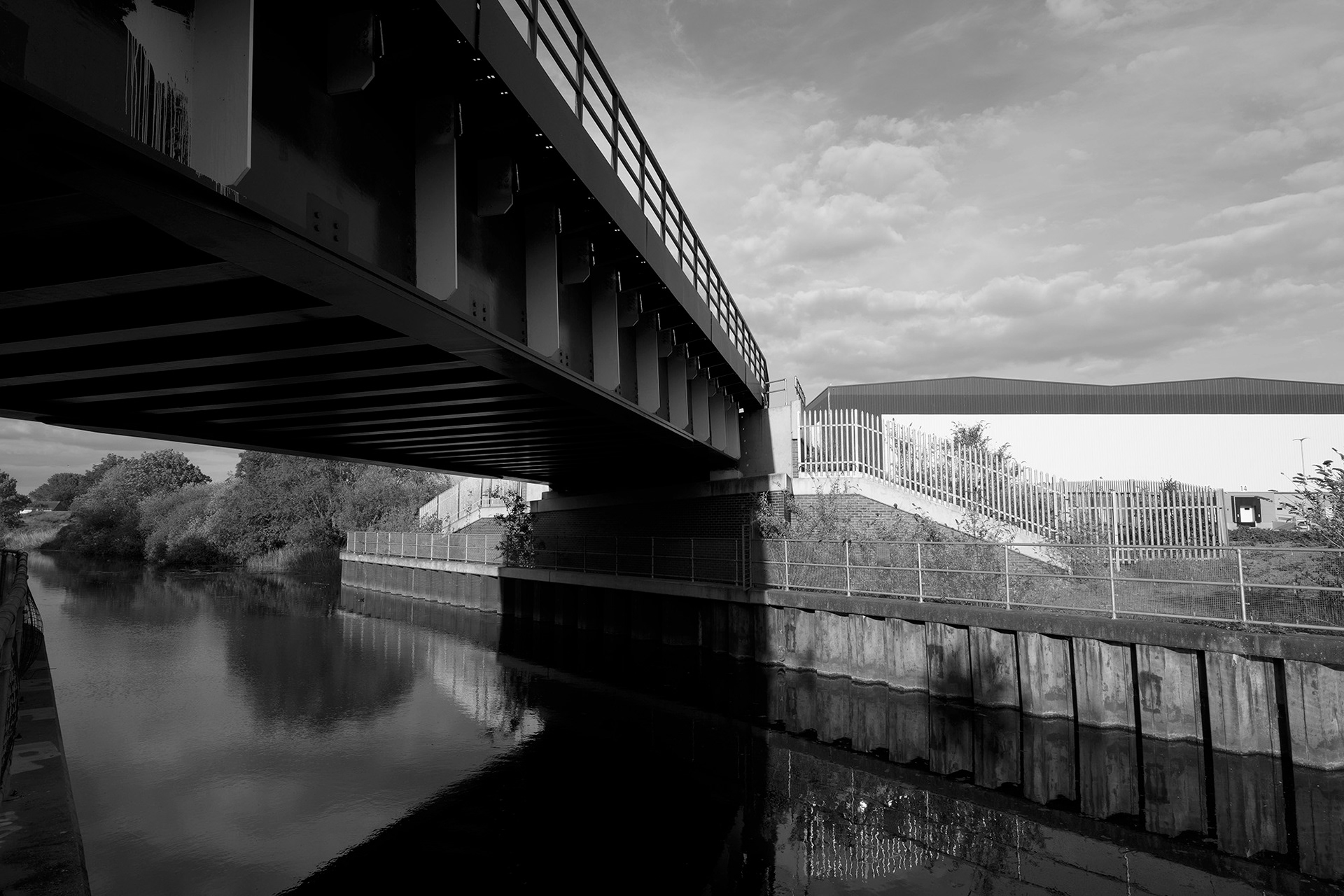 A black and white photo of a river in an industrial setting with a metal bridge.