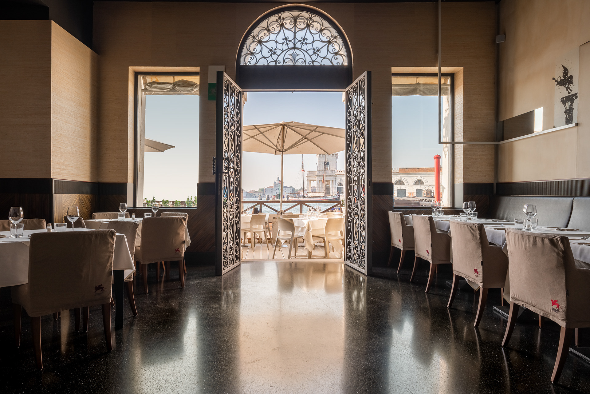 In questa suggestiva immagine di Joan Porcel, ci immergiamo nell'atmosfera avvolgente dell'interno del ristorante "Biennale" sul Canal Grande, di fronte alla Basilica della Salute. La scena racconta una storia di raffinatezza veneziana, combinando sapientemente elementi tradizionali e moderni.  La vista mozzafiato sul Canal Grande è il cuore dell'esperienza, con ampie finestre che permettono alla luce naturale di danzare sulle acque, creando un'atmosfera di lusso e serenità. La Basilica della Salute fa da sfondo, aggiungendo una dimensione di storia e grandezza alla scena.  La disposizione degli arredi riflette un'eleganza senza sforzo, con tavoli ben apparecchiati e sedie che invitano gli ospiti a godere di una cucina prelibata in questo ambiente straordinario. I dettagli veneziani, come il vetro soffiato e i tessuti riccamente intessuti, aggiungono un tocco di autenticità locale.  Joan Porcel cattura la magia di questo ristorante veneziano con la sua abilità nell'immortalare dettagli significativi. Ogni elemento, dalle decorazioni murali agli arredi attentamente selezionati, sembra narrare la storia di un luogo che incarna l'eleganza e il fascino unici di Venezia.  English:  In this captivating image by Joan Porcel, we are immersed in the enveloping atmosphere of the interior of the "Biennale" restaurant on the Grand Canal, in front of the Basilica della Salute. The scene tells a story of Venetian sophistication, skillfully combining traditional and modern elements.  The breathtaking view of the Grand Canal is the centerpiece of the experience, with expansive windows allowing natural light to dance on the waters, creating an atmosphere of luxury and serenity. The Basilica della Salute serves as a backdrop, adding a dimension of history and grandeur to the scene.  The arrangement of furnishings reflects effortless elegance, with well-set tables and chairs inviting guests to enjoy exquisite cuisine in this extraordinary setting. Venetian details, such as blown glass and richly woven fabrics, add a touch of local authenticity.  Joan Porcel captures the magic of this Venetian restaurant with his skill in immortalizing significant details. Every element, from mural decorations to carefully selected furnishings, seems to narrate the story of a place that embodies the unique elegance and charm of Venice.