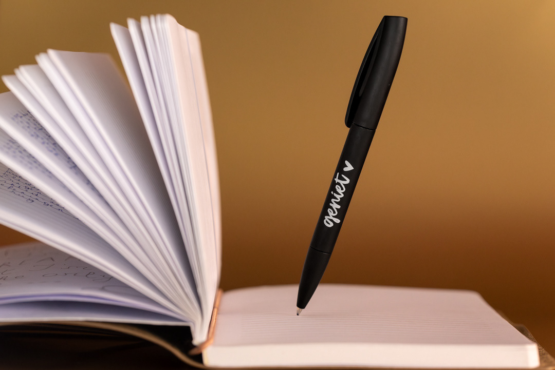 Een balpen met het opschrift 'geniet' zweeft boven een open schrift.