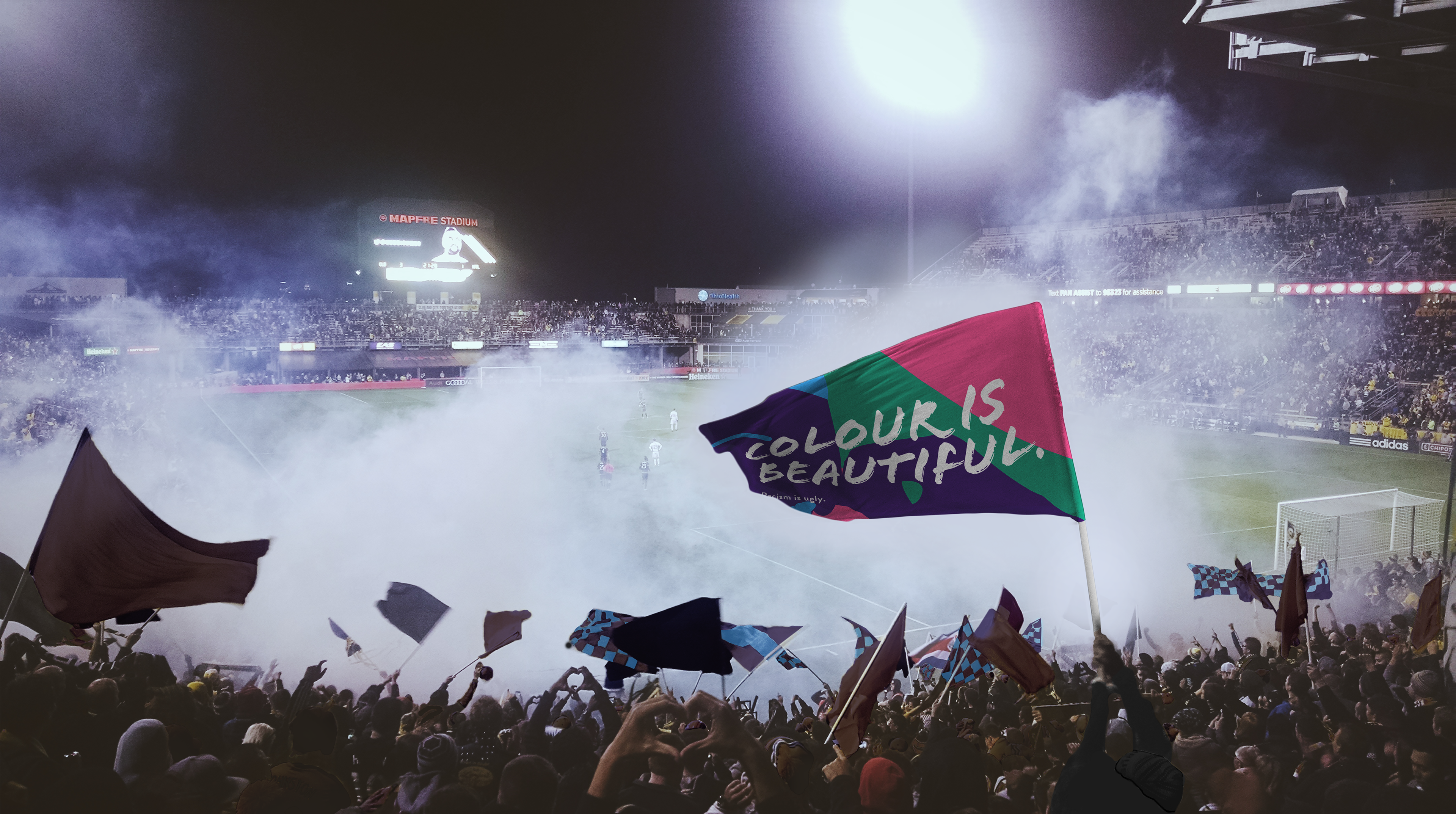 A stadium with the tifo ultras section of fans with on of them holding a flag with the Colour is Beautiful branding on it.