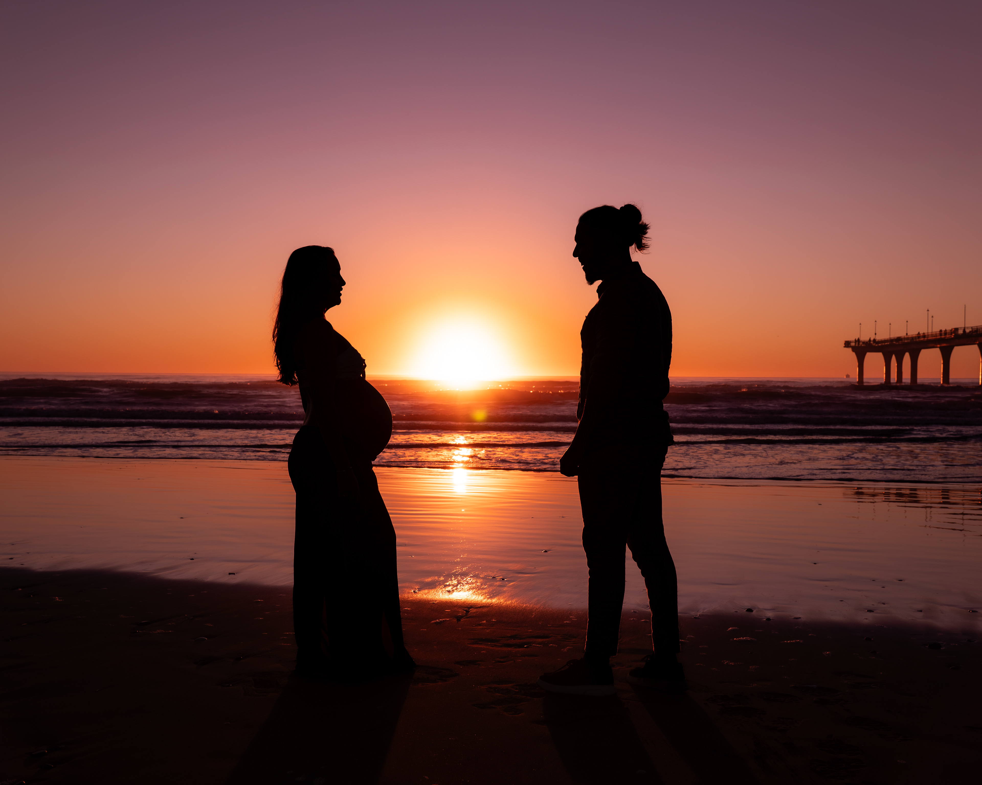 Silhouette of a pregnant woman and a man facing each other on a beach at sunset, with the sun reflecting on the ocean and a pier visible in the background.