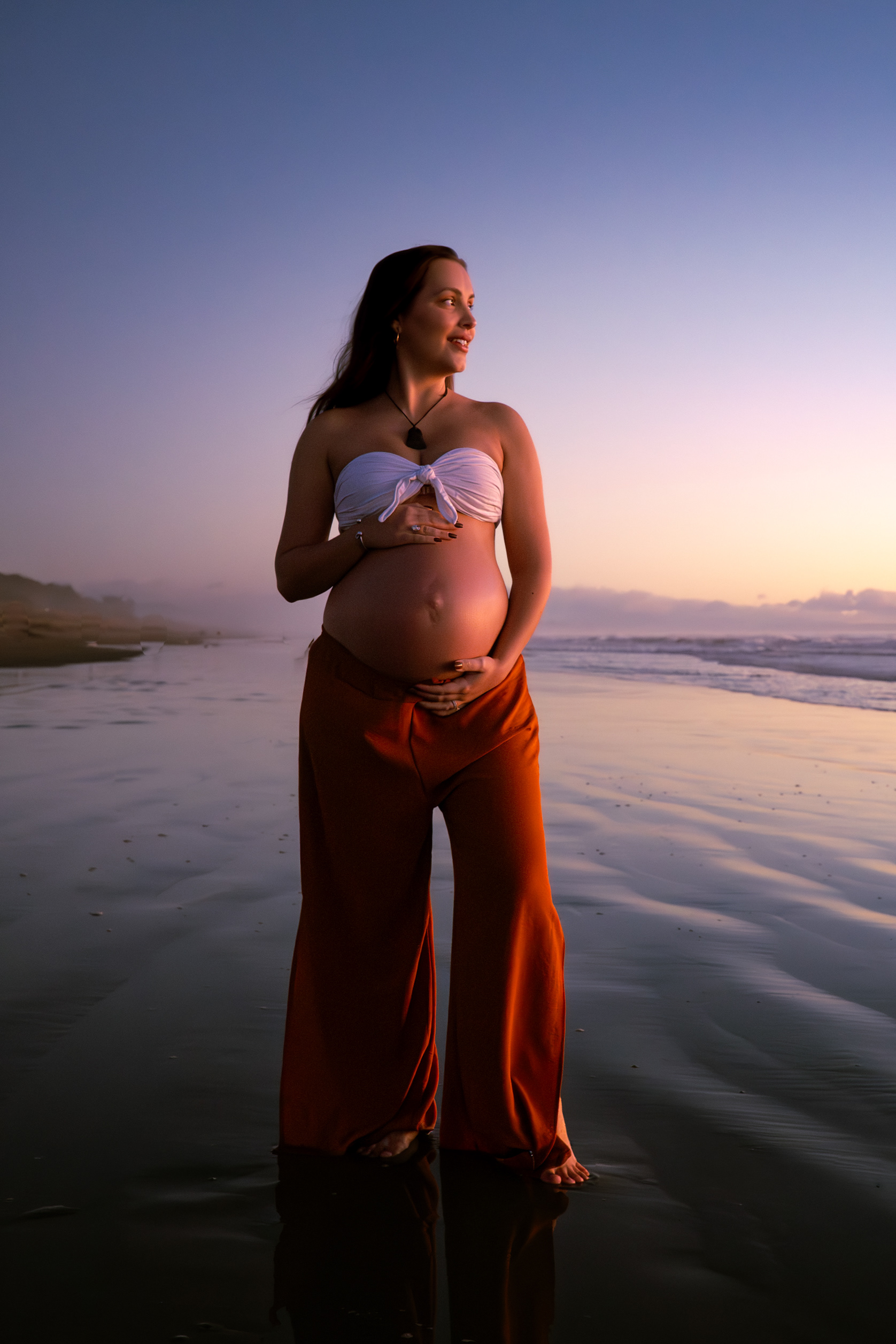 A pregnant woman in an orange pantsuit and white top standing on the beach at sunset, looking out to sea.