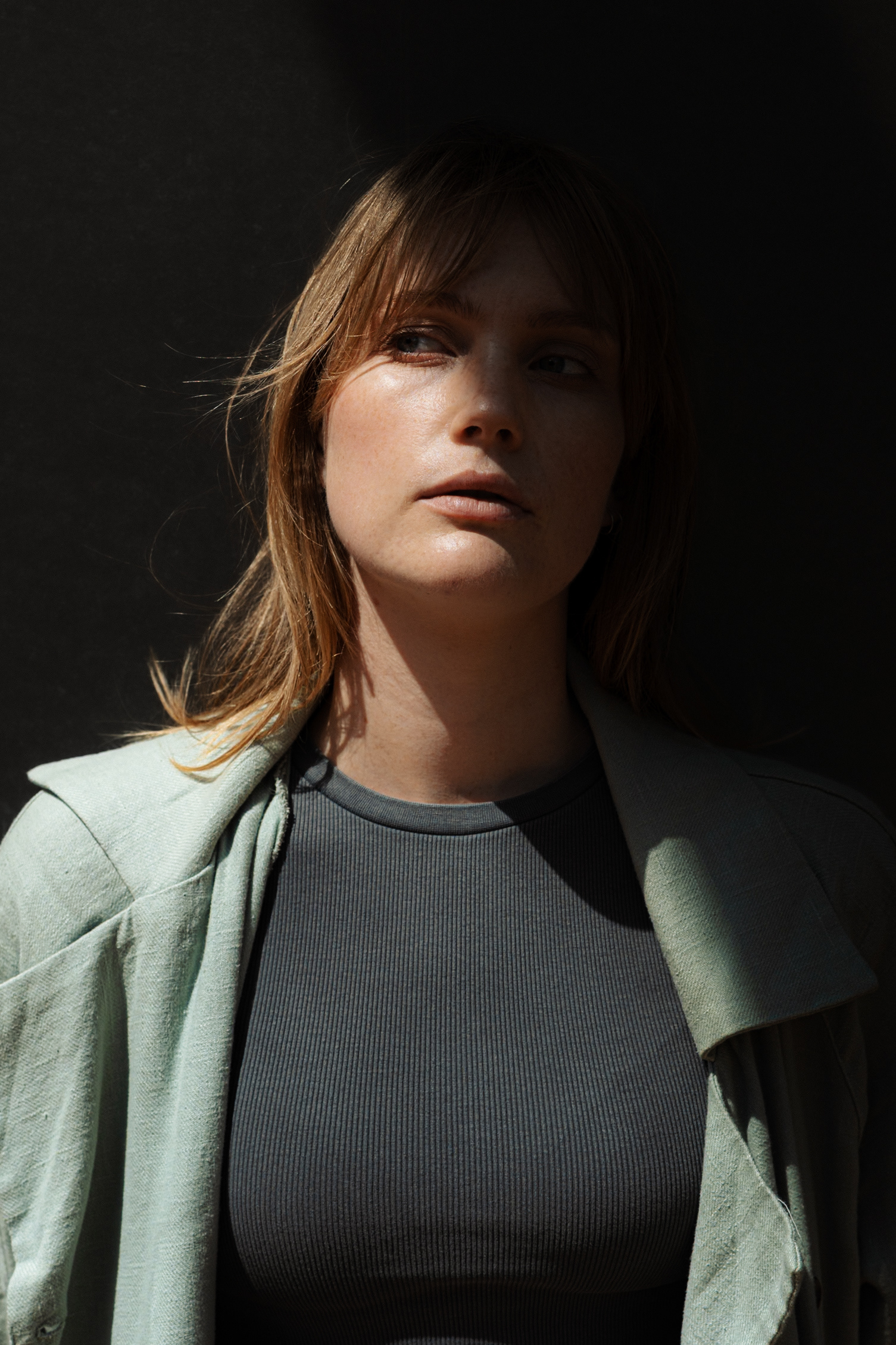 A close-up portrait of a woman with blonde bangs and a light green jacket, illuminated by sharp sunlight against a dark background