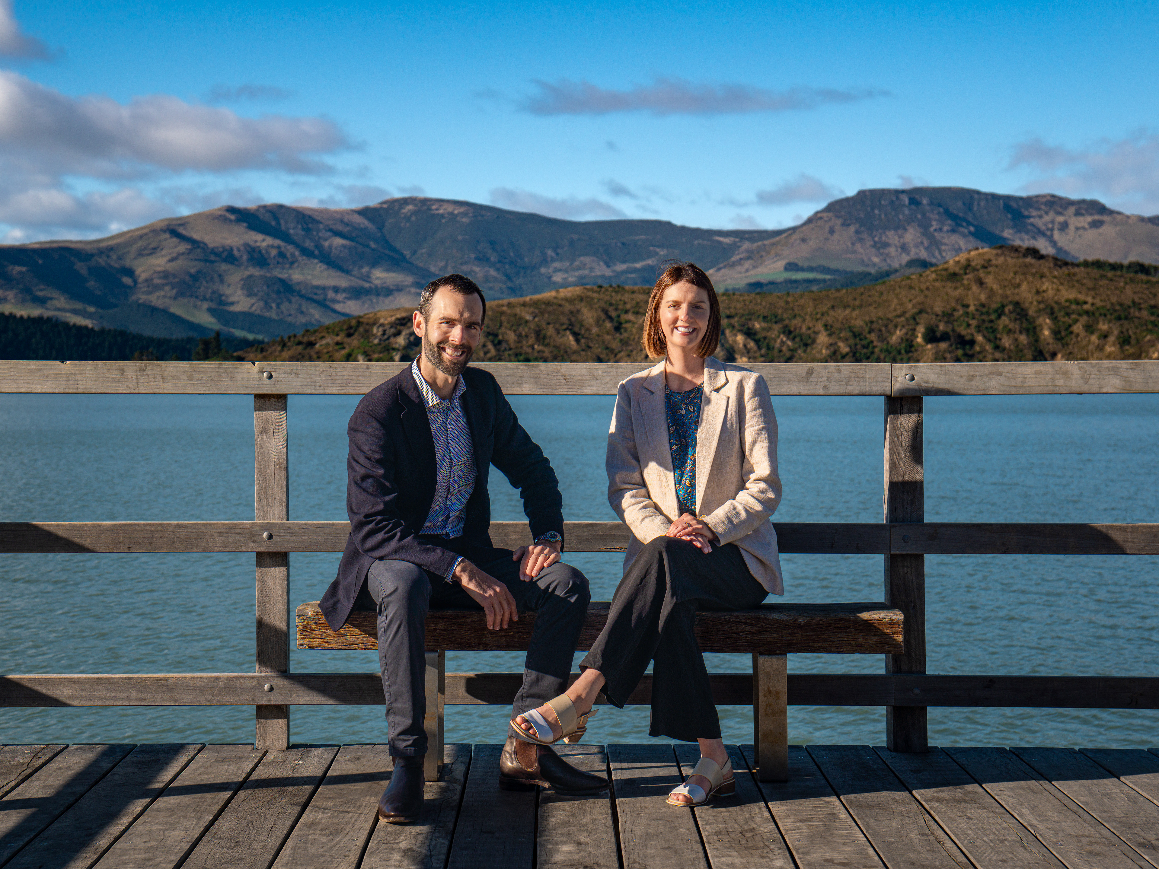 A smiling man and woman sitting on a wooden bench on a pier, with a calm body of water and rolling hills under a blue sky in the background.