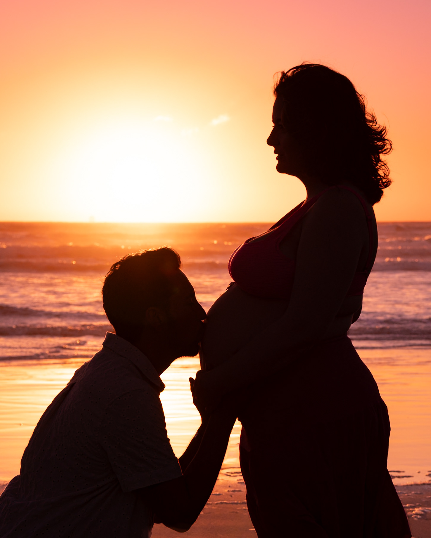A silhouette of a couple on the beach at sunset, with the man kneeling and kissing the woman's pregnant belly.
