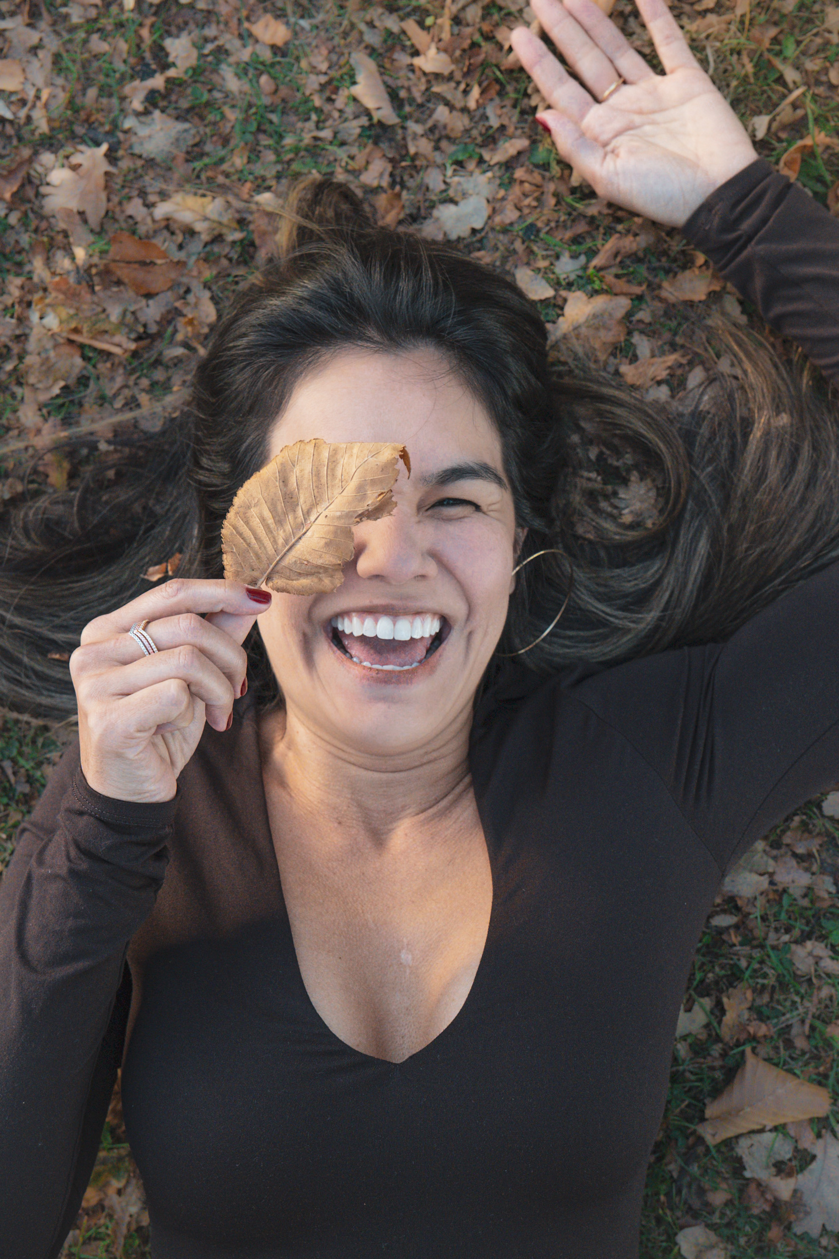 A top-down portrait of a smiling woman lying on autumn leaves, playfully holding a dry brown leaf over one eye.