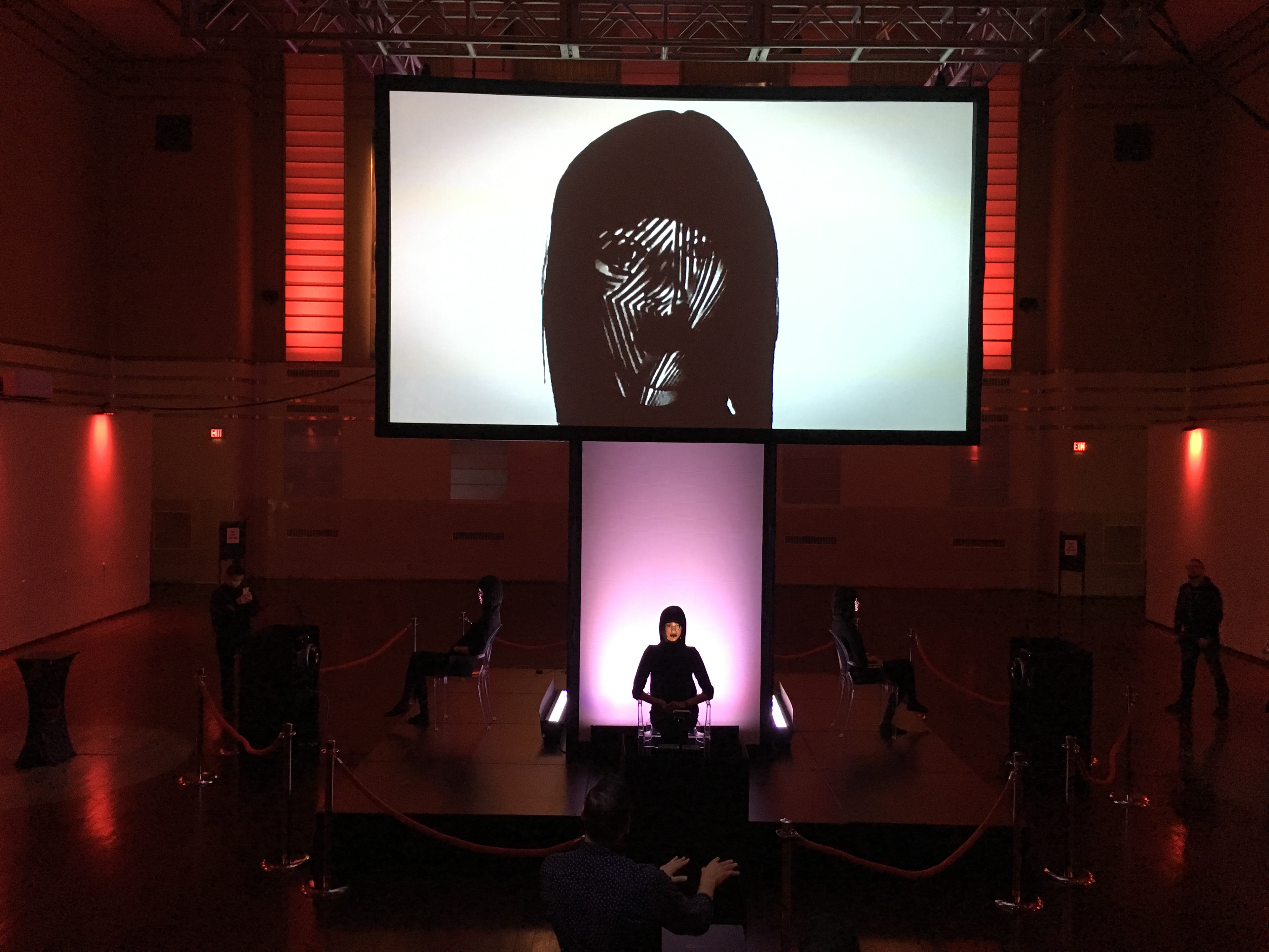 A model sits on a stage with her face projected onto a large screen above her.
