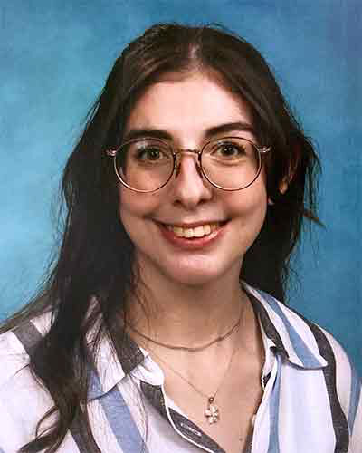 Photo of Nicole, a white woman in her late twenties with medium-length brown hair and glasses. She is smiling, wearing a white and blue striped button-up shirt, and sitting in front of a blue background.