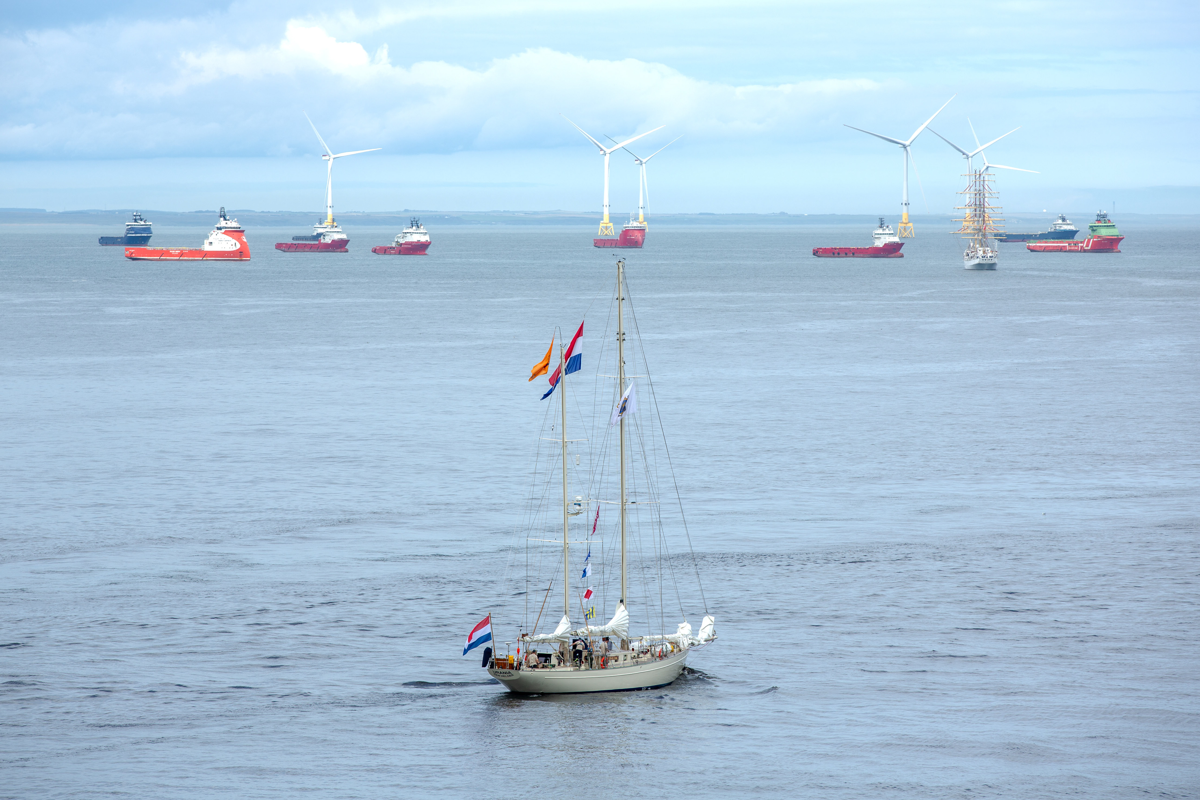 A lone tall ship slips past Aberdeen’s offshore wind turbines, sails meeting steel, while oilfield supply vessels watch on. Old routes and new energies,