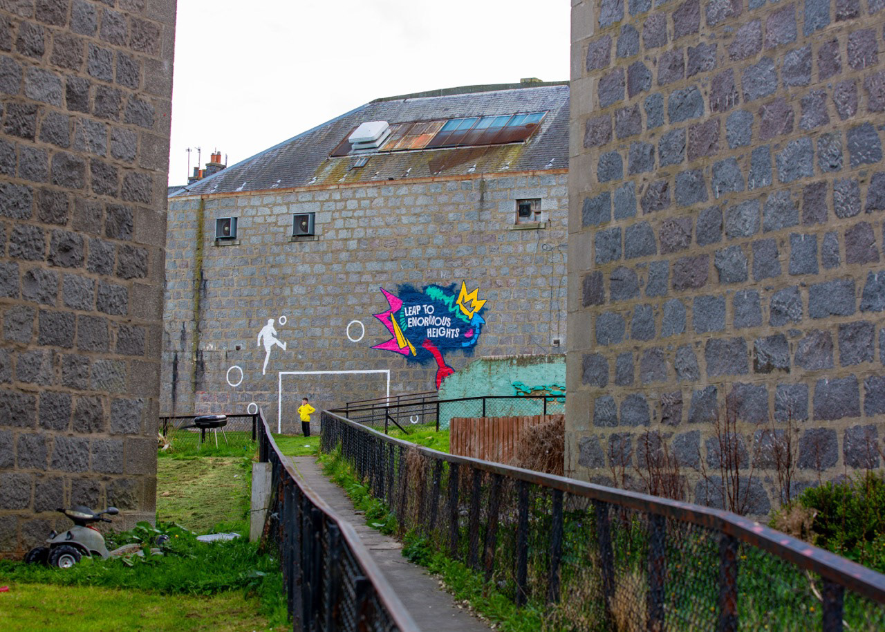 Leap to Enormous Heights finished and alive with play. A local Printfield lad steps into goal beneath the completed mural, turning public art into a lived moment of pride and possibility