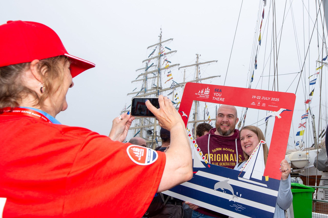 A brief pause amid the bustle — a young couple smile as a tall ship volunteer captures the moment, a small keepsake from a shared day by the harbour