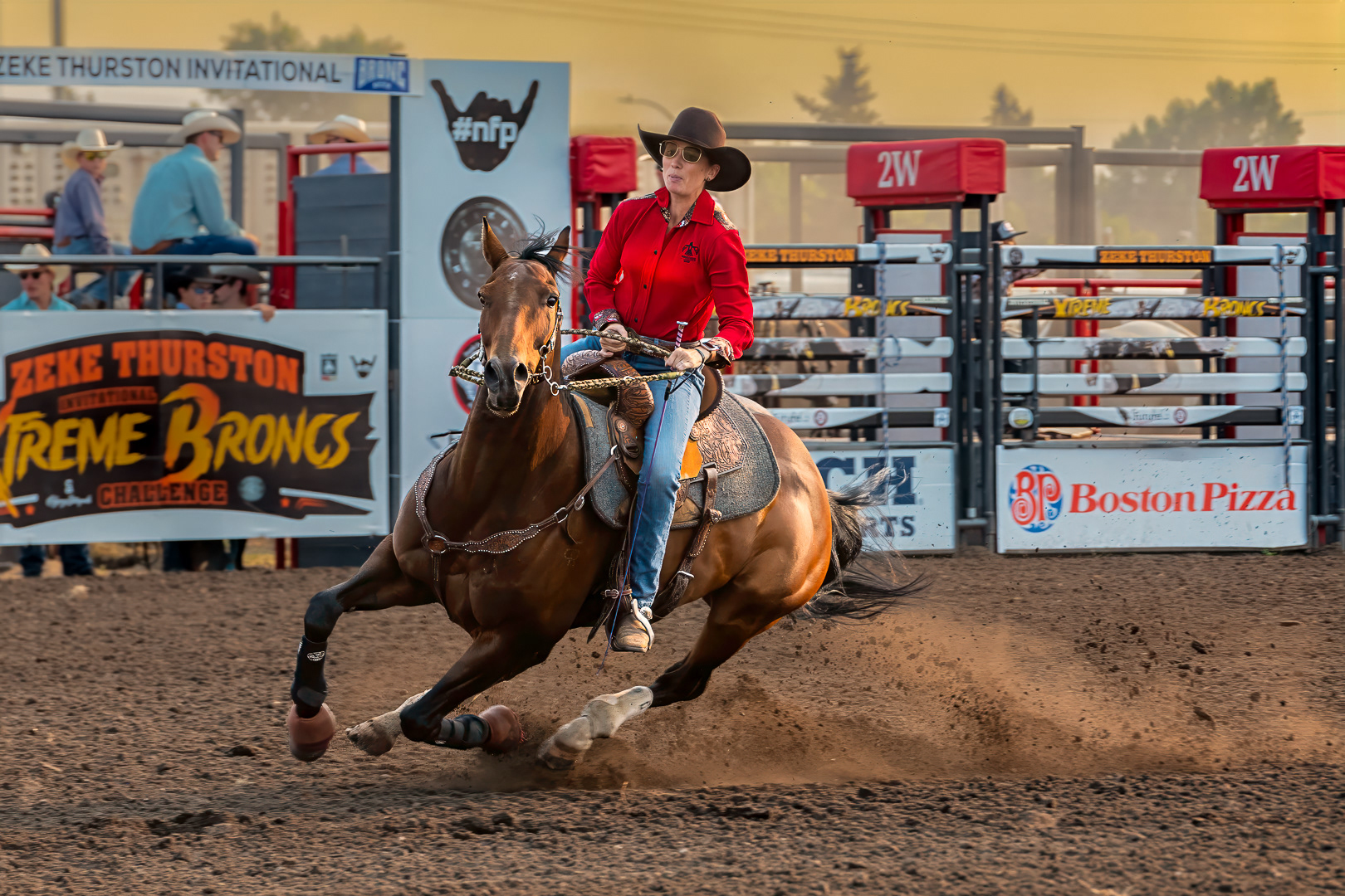 A cowgirl wearing a red long-sleeve shirt and cowboy hat makes a sharp, fast turn on a brown horse during a rodeo event, kicking up dirt in an arena framed by arena banners and bucking chutes. A rodeo rider wearing a dark cowboy hat, sunglasses, a red long-sleeved button-up shirt, and blue jeans leans into a sharp, high-speed turn on a brown horse. The horse is angled steeply toward the left, kicking up a plume of brown dirt across the arena floor. In the background are metal rodeo chutes with red padding labeled "2W," sponsor banners including "Zeke Thurston Invitational Xtreme Broncs Challenge" and "Boston Pizza," and spectators watching behind the arena fence under warm outdoor light.