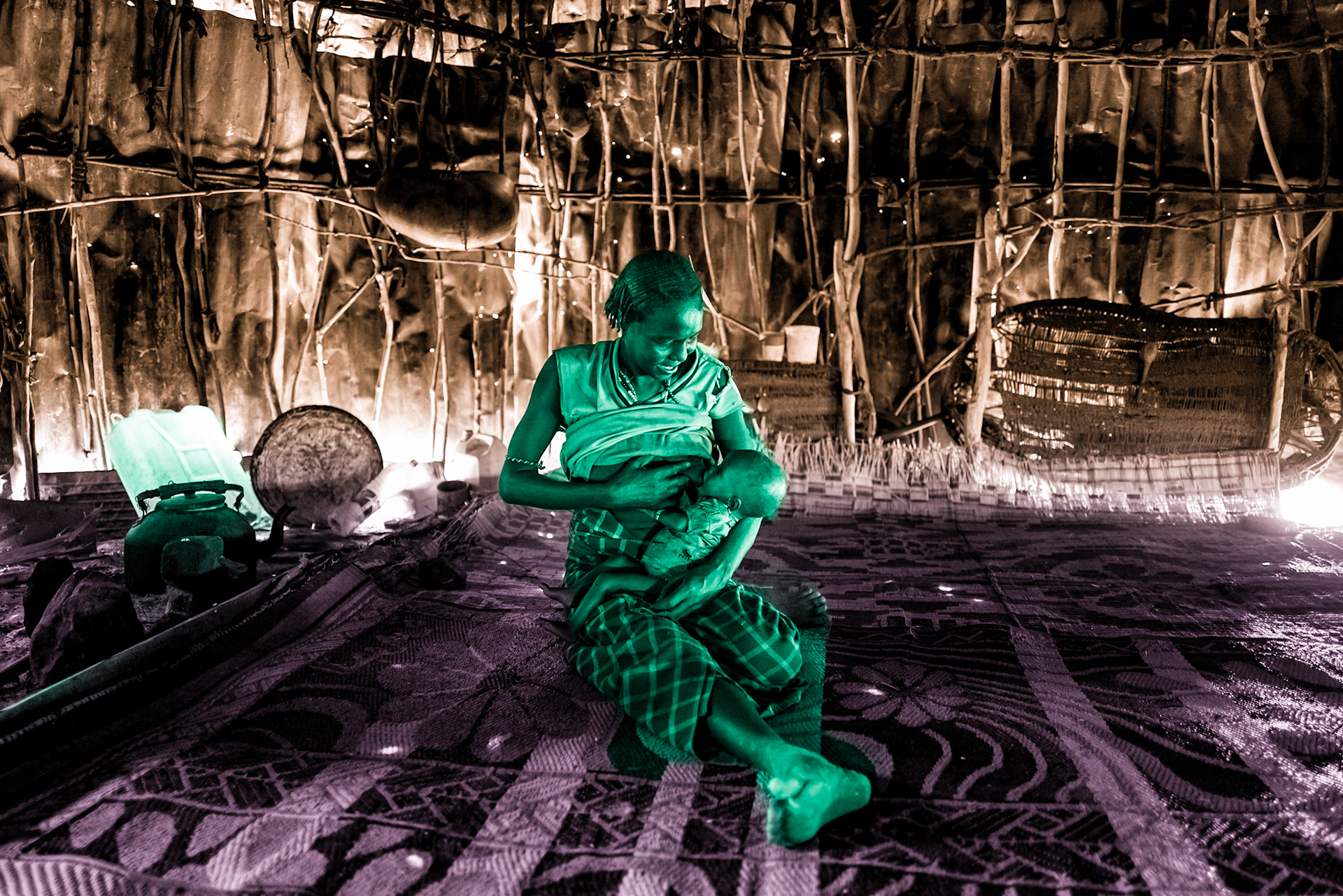 A Daasanach mother is nursing her baby inside a hut made of iron sheets and wooden sticks. The region is experiencing a major disaster due to both armed conflict and a severe drought, which has caused crop failures and livestock deaths, leading to the worst famine in East Africa in decades.