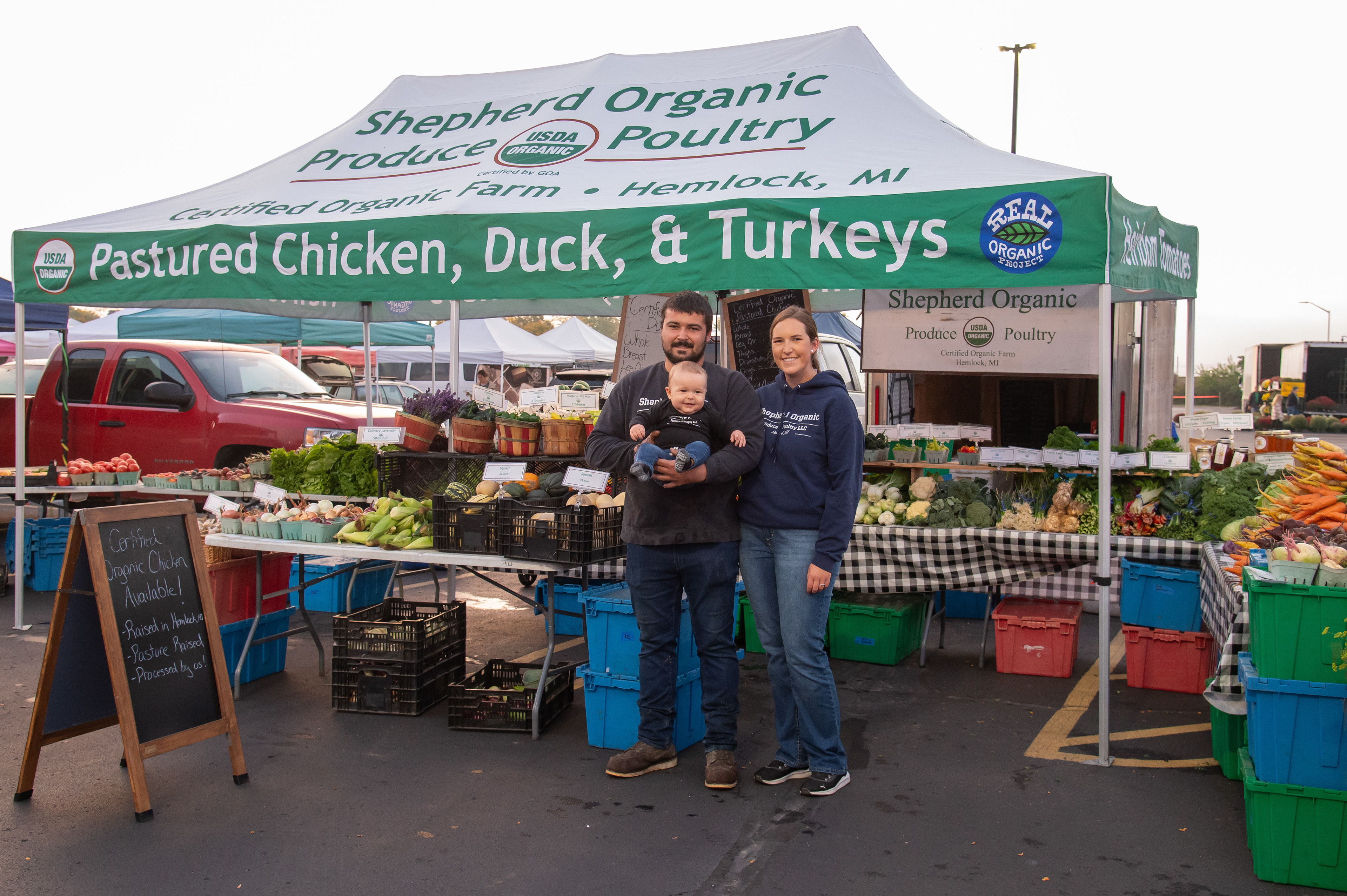 Tyler and Hannah Shepherd pose with their son Adam in front of their stand at the Midland Farmer’s Market, 825 East Main Street, Midland, Michigan, Saturday, September 30, 2023. With most workdays starting around six in the morning, Tyler said a lot of his time on the farm is spent picking. On some market days, he is up as early as 2:45 a.m. to load his truck and trailer.